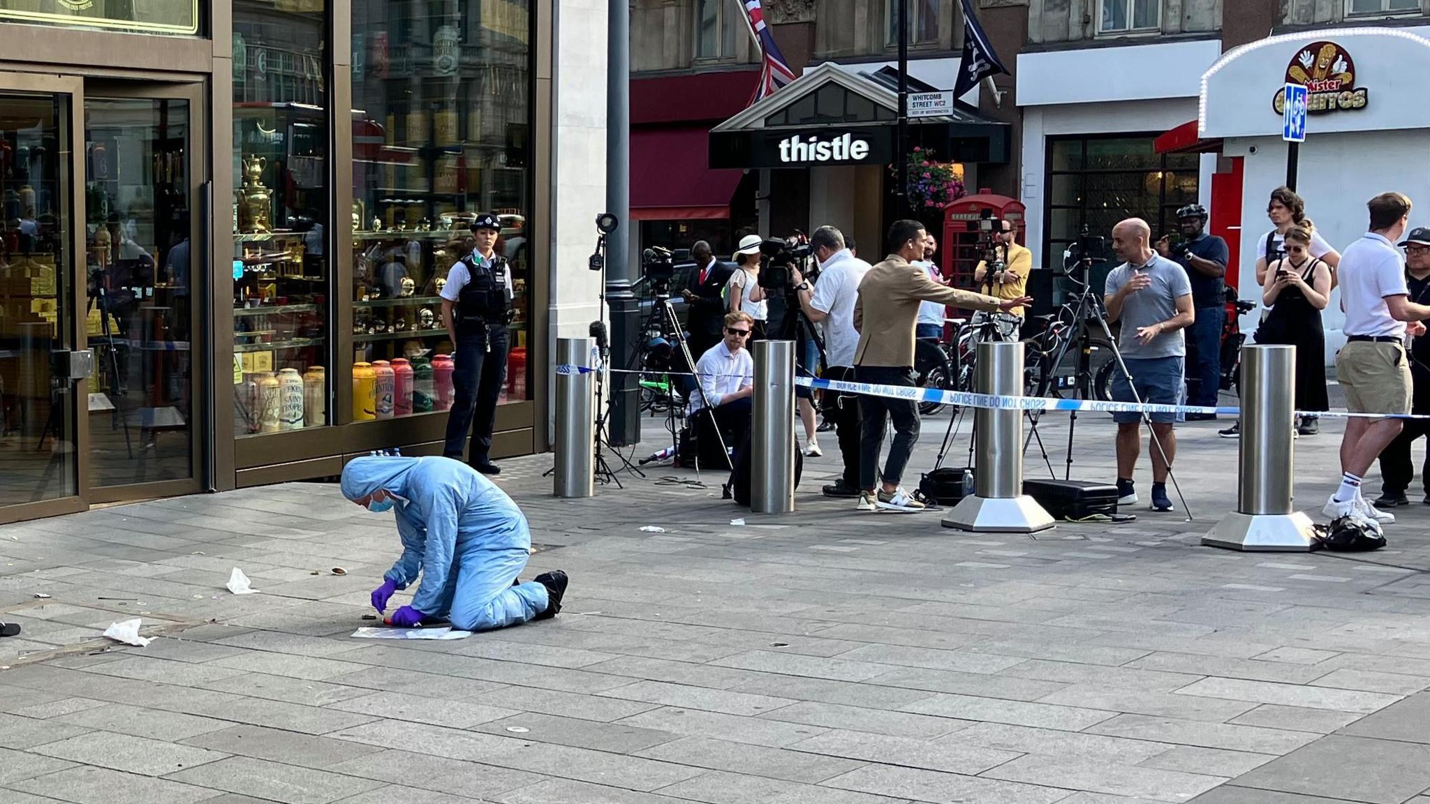 Forensic officer wearing blue covering and face mask bends over on pavement outside shop with a police officer and camera crews behind them