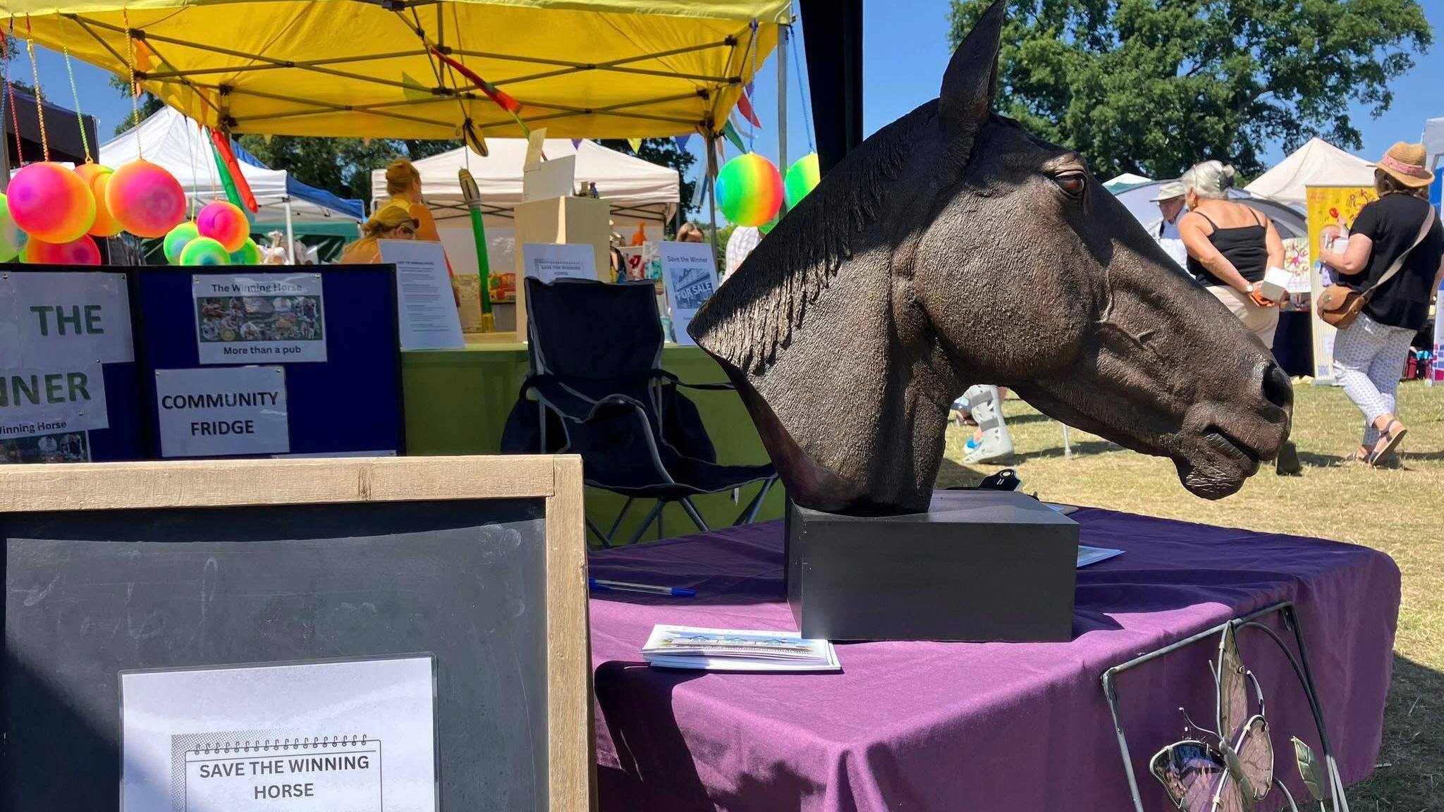 A brass sculpture of a horse's head sits on a table covered with a purple cloth at a community event, with publicity literature from the Save the Winner campaign scattered around and pinned up on boards around it.