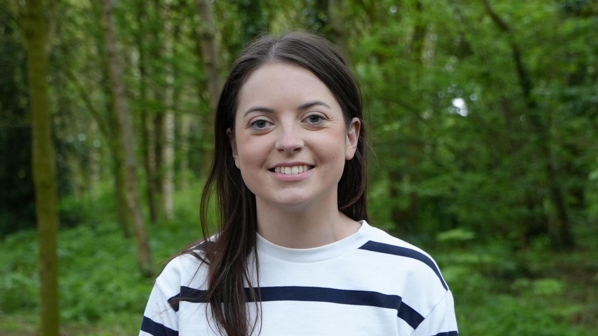 Kathryn is stood in a wooded area, she is wearing a white striped jumper and has long, dark hair.
