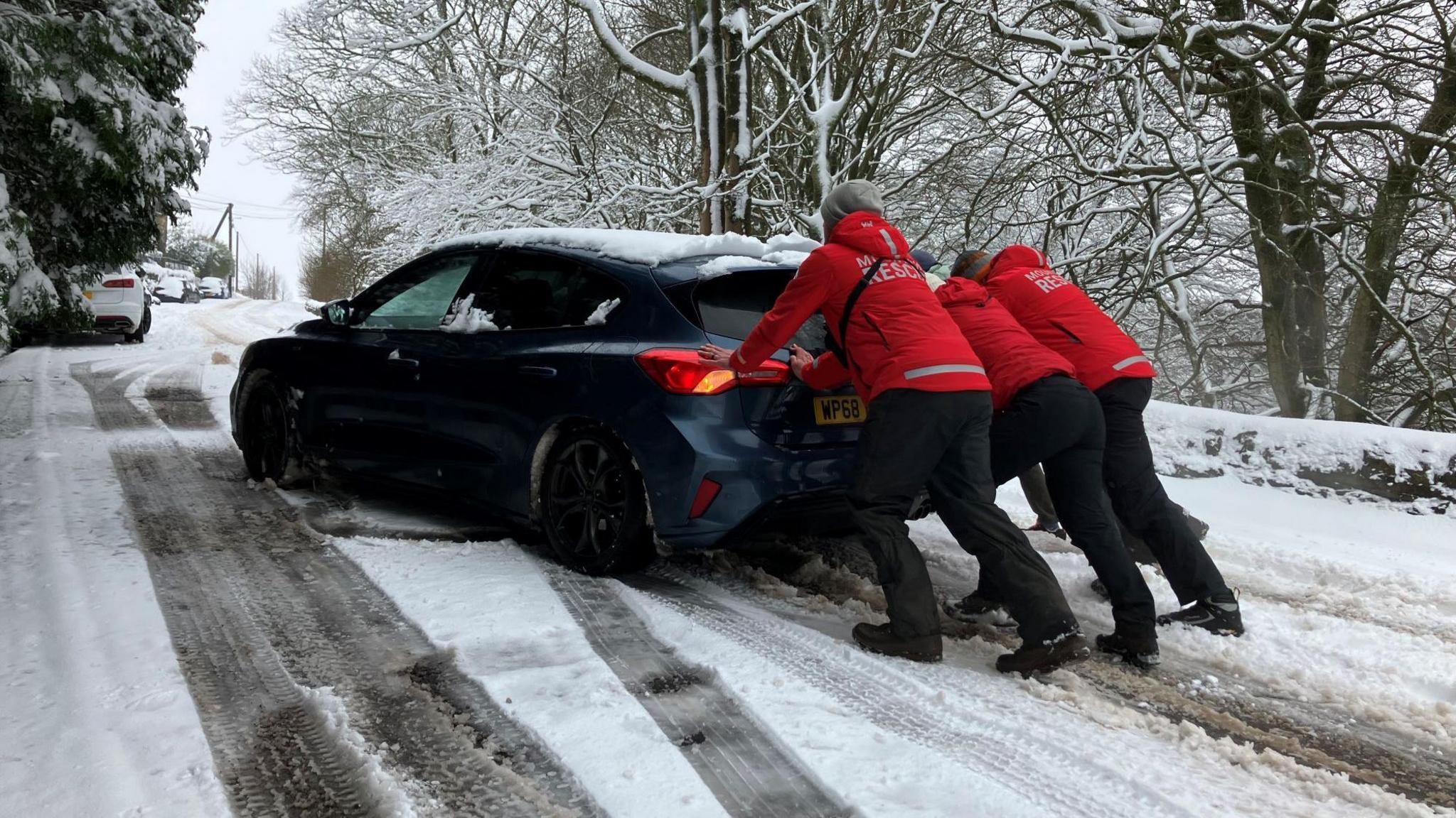 Three people in red mountain rescue jackets help to push a blue car up a snowy road. 