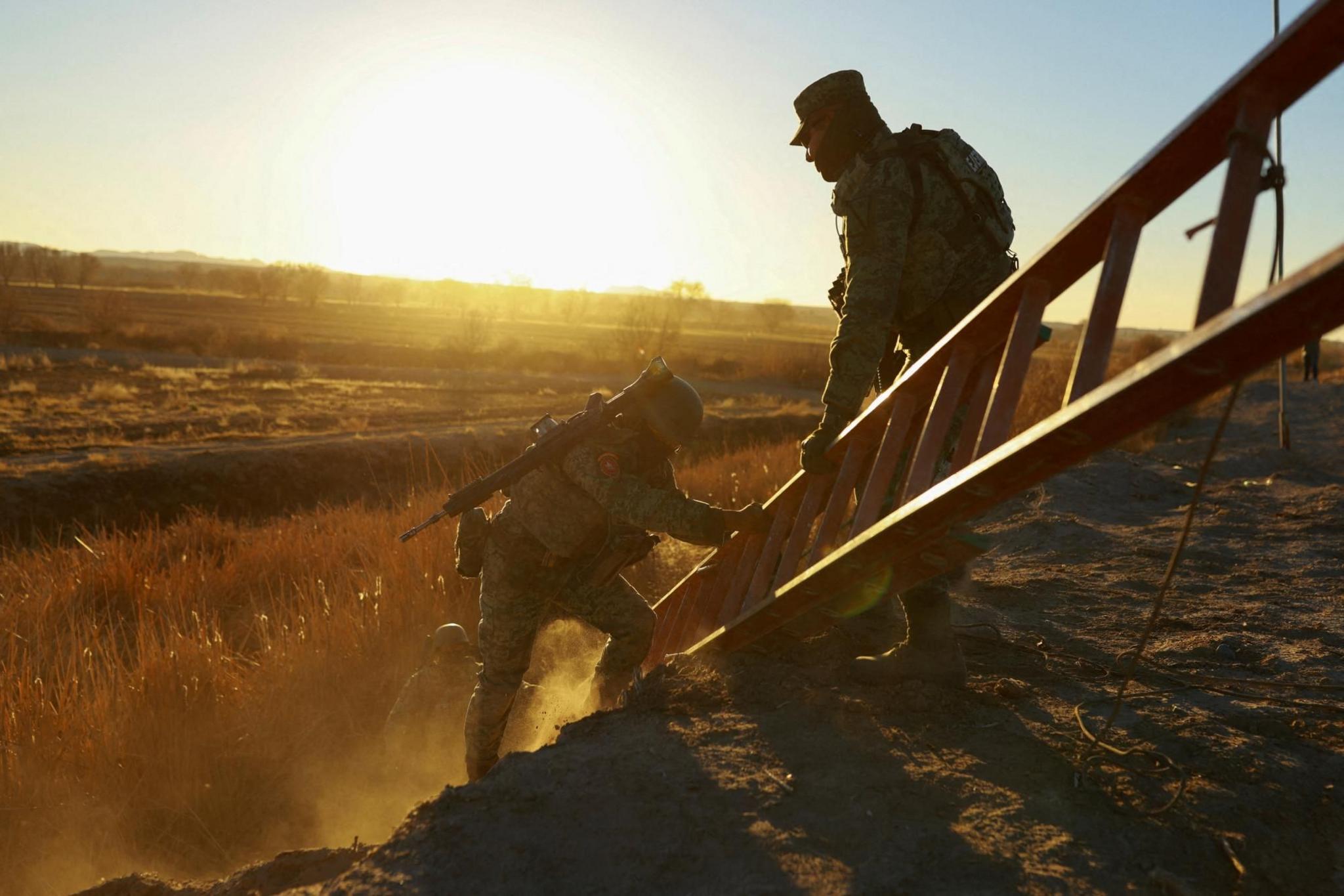 Two members of the Mexican National Guard lift a ladder up a slope. One of the guards is holding a rifle. The sun is beating down in the background.