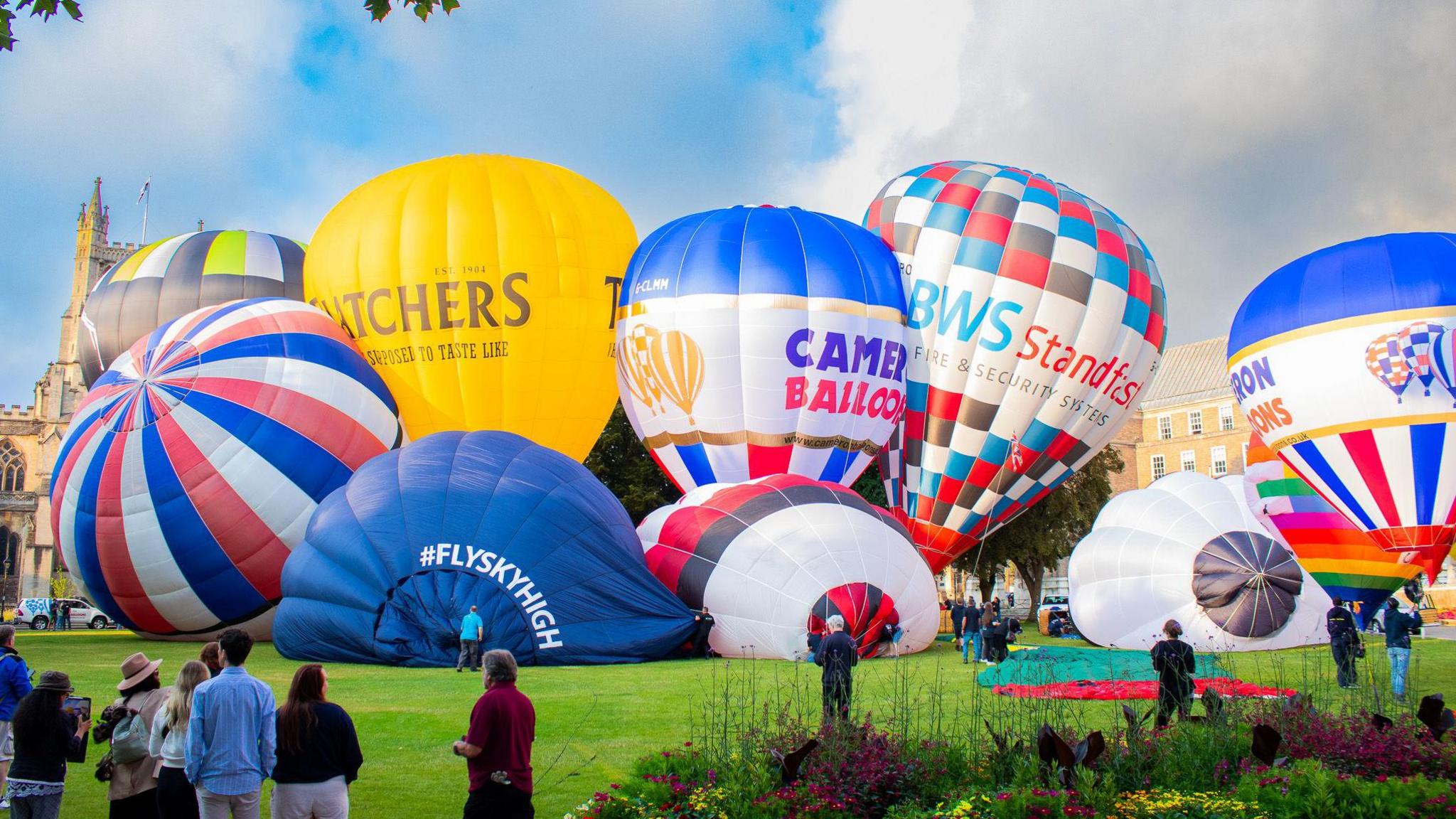 A selection of about nine hot air balloons on the ground in various states of deflation. They are all colourful with bright yellow, red and blue designs. You can see the top of Bristol Cathedral in the background and some people observing the balloons in the foreground.