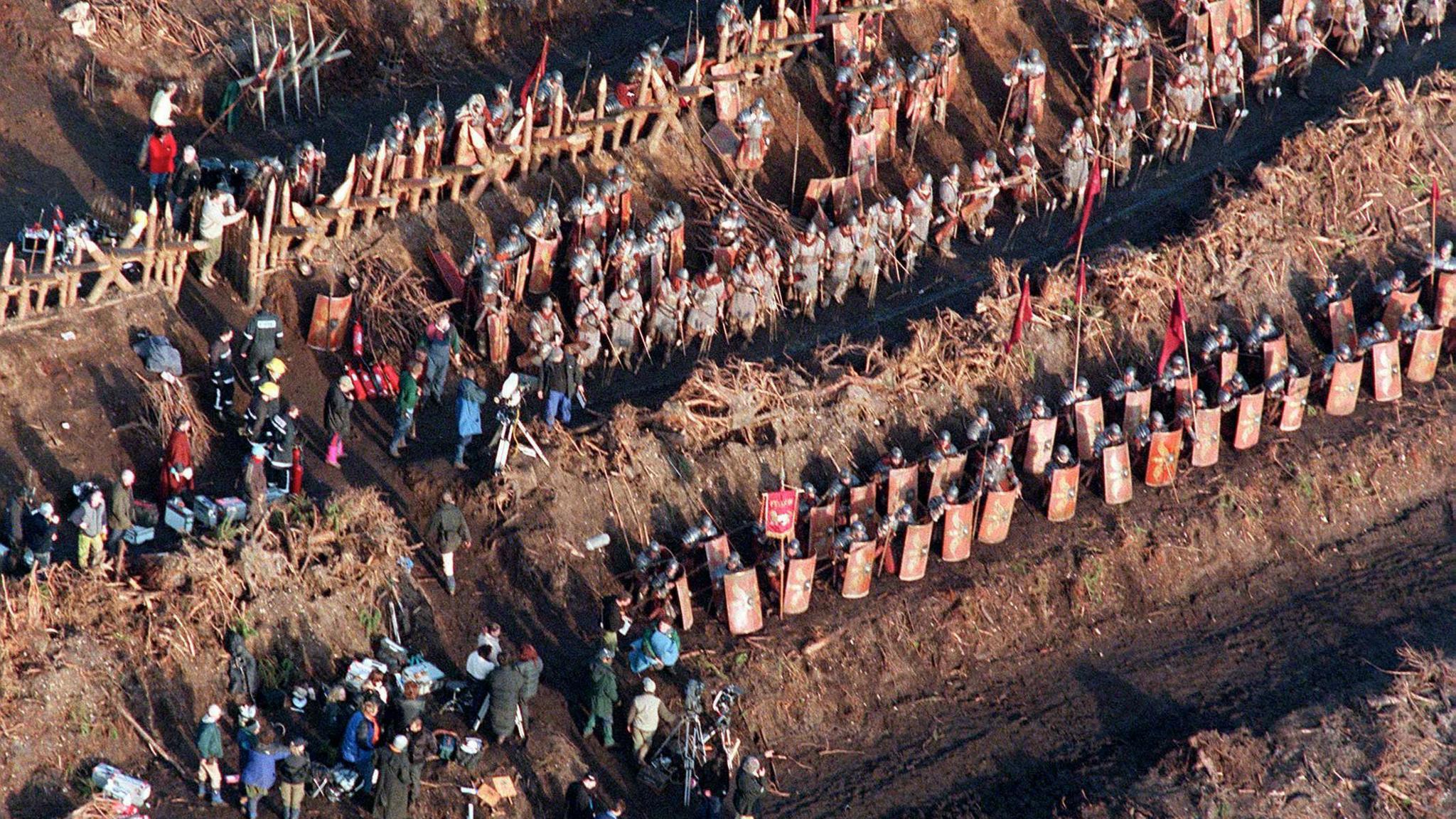 An aerial shot of rows of extras dressed as soldiers, carrying swords and shields, with a large filming crew to the side of the field.