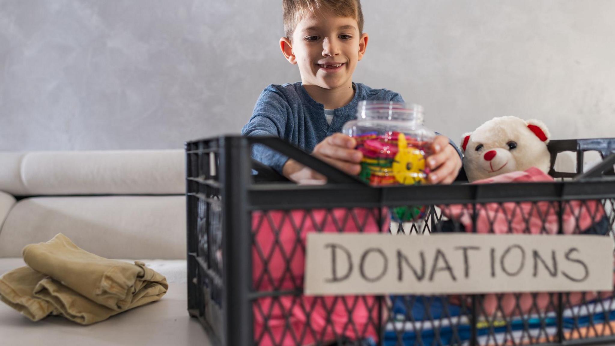 Young boy packing clothes, toys and a teddy bear in a donation box.