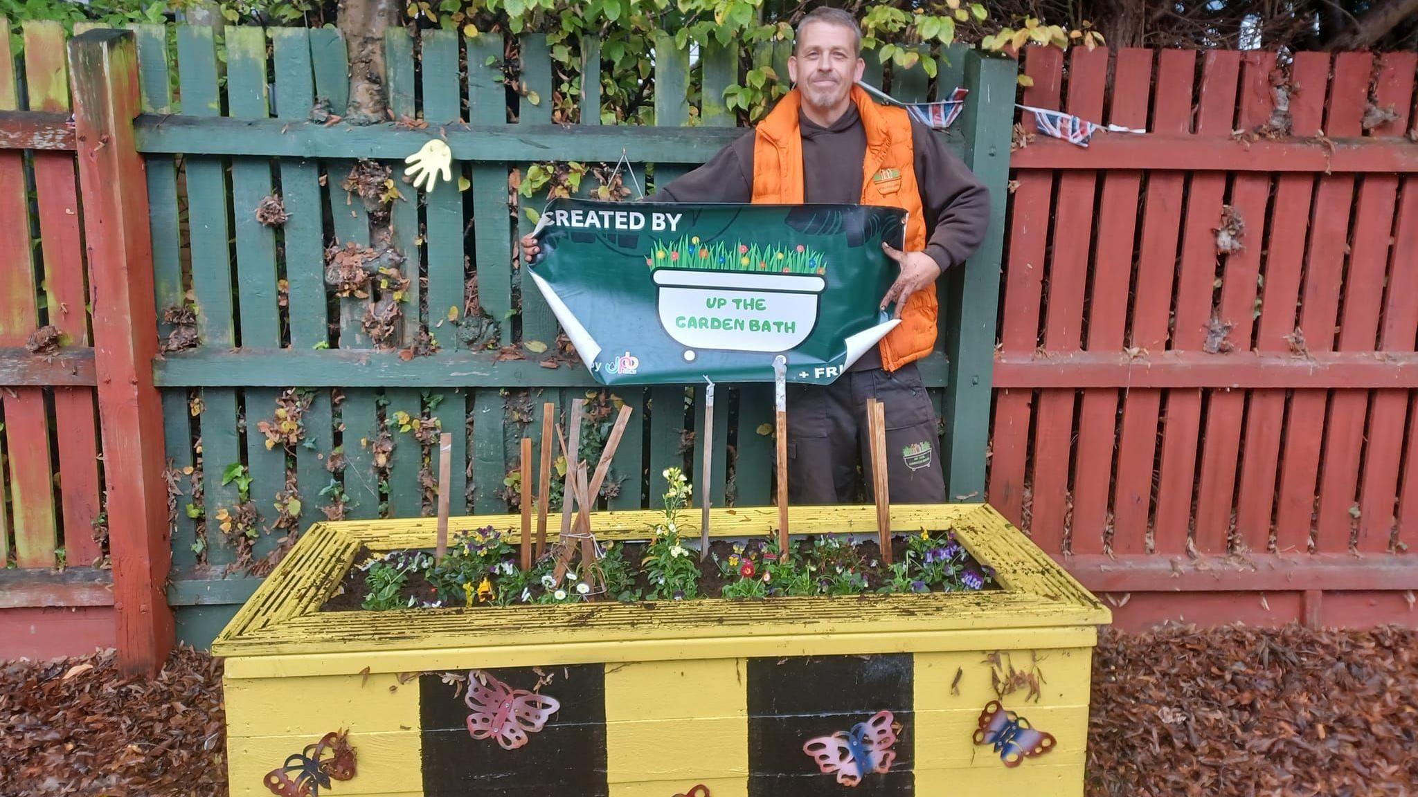 Dave Poulton is stood outside behind an upcycled bathtub, which has been covered in wood to make it look like a planter. It is black and yellow, striped like a bee, with butterflies sprayed on the side. Dave is wearing an orange gillet and is holding a "Created by Up the Garden Bath" sign in his hands. 