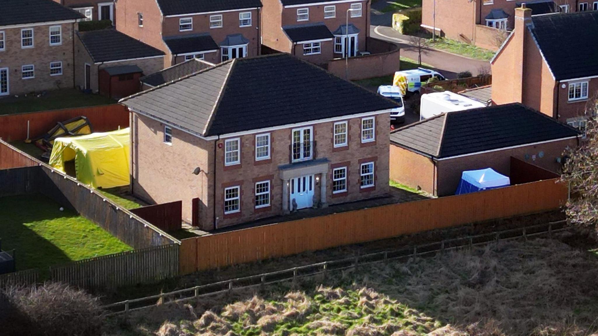 Drone shot of a modern large detached house, it has two storeys and a garden and garage next to it with a large yellow police forensic tent in the garden