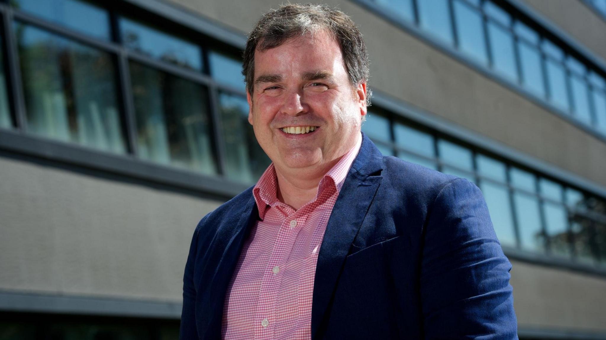 Renfrewshire Council leader Iain Nicolson, who has short greying hair and is wearing a pink check shirt with a navy blue blazer, stands smiling outside Renfrewshire House - a modern building with rows of black-framed windows..