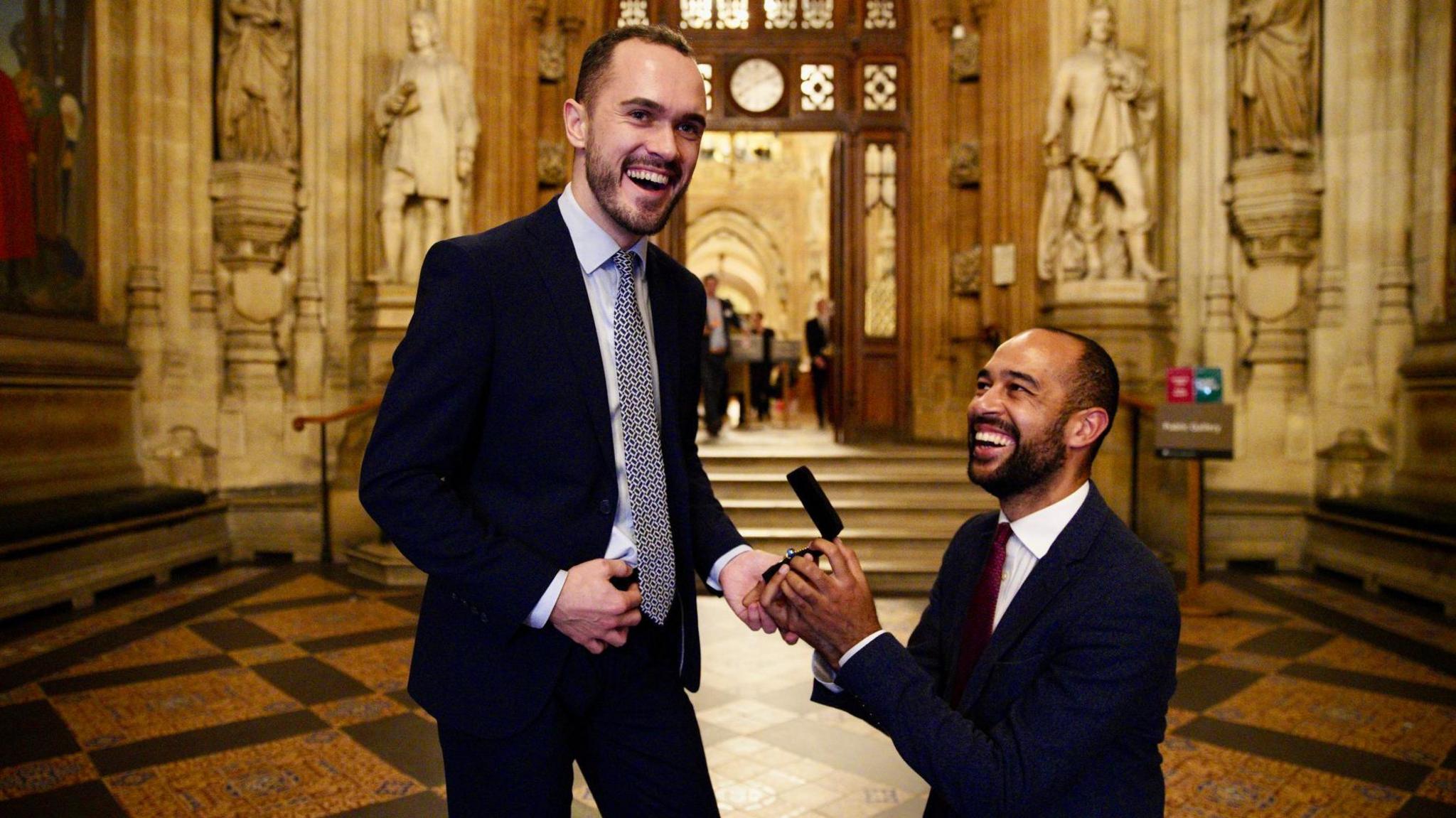 A picture of Eastbourne MP Josh Babarinde and his partner Connor at the House of Commons in London. Josh is on his knees holding an engagement ring for Connor, who is stood up. Both are smiling and wearing navy suits.