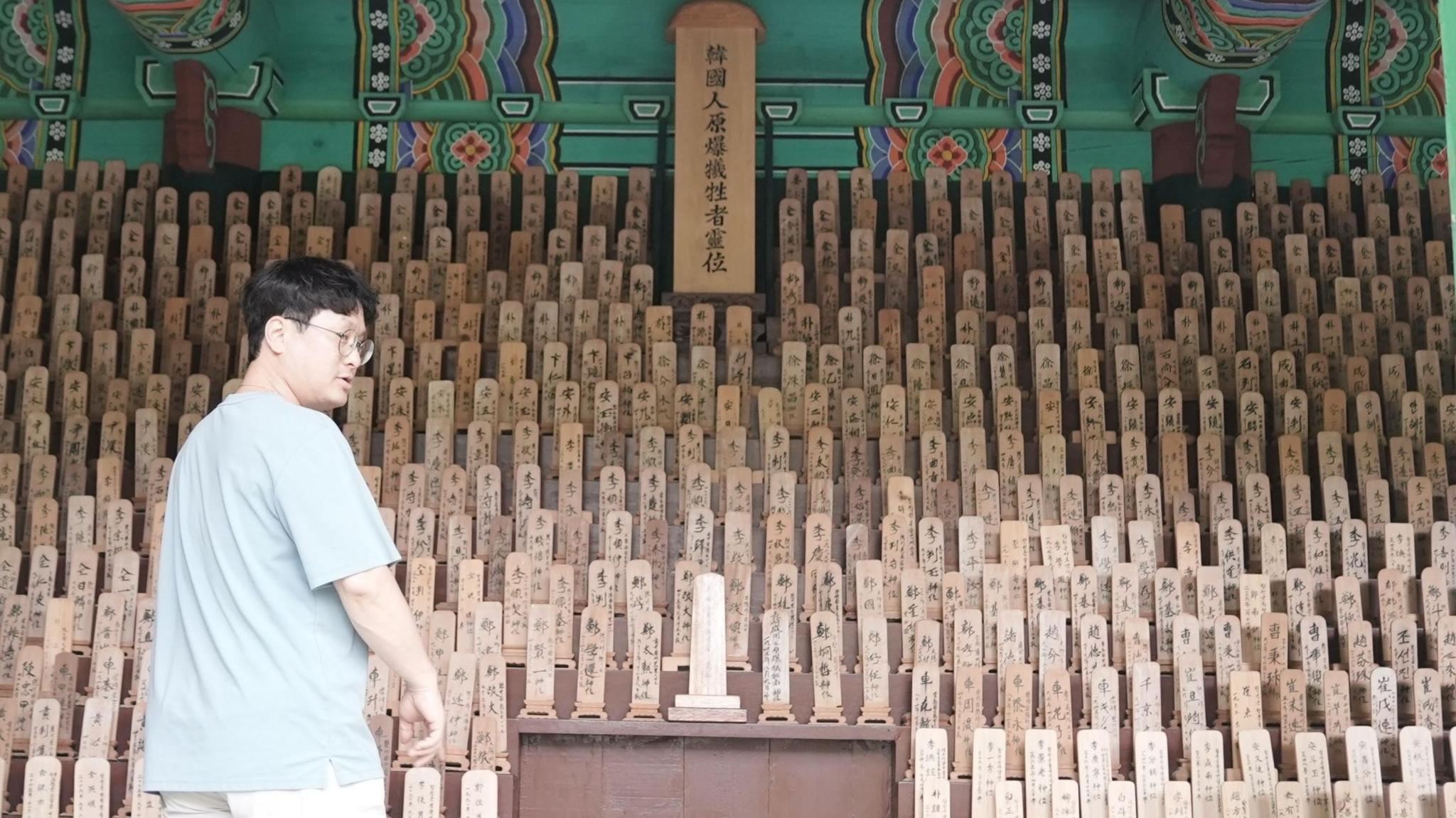 A man stands at a temple in front of rows of small rectangular wooden planks, standing vertically, with Chinese characters on them