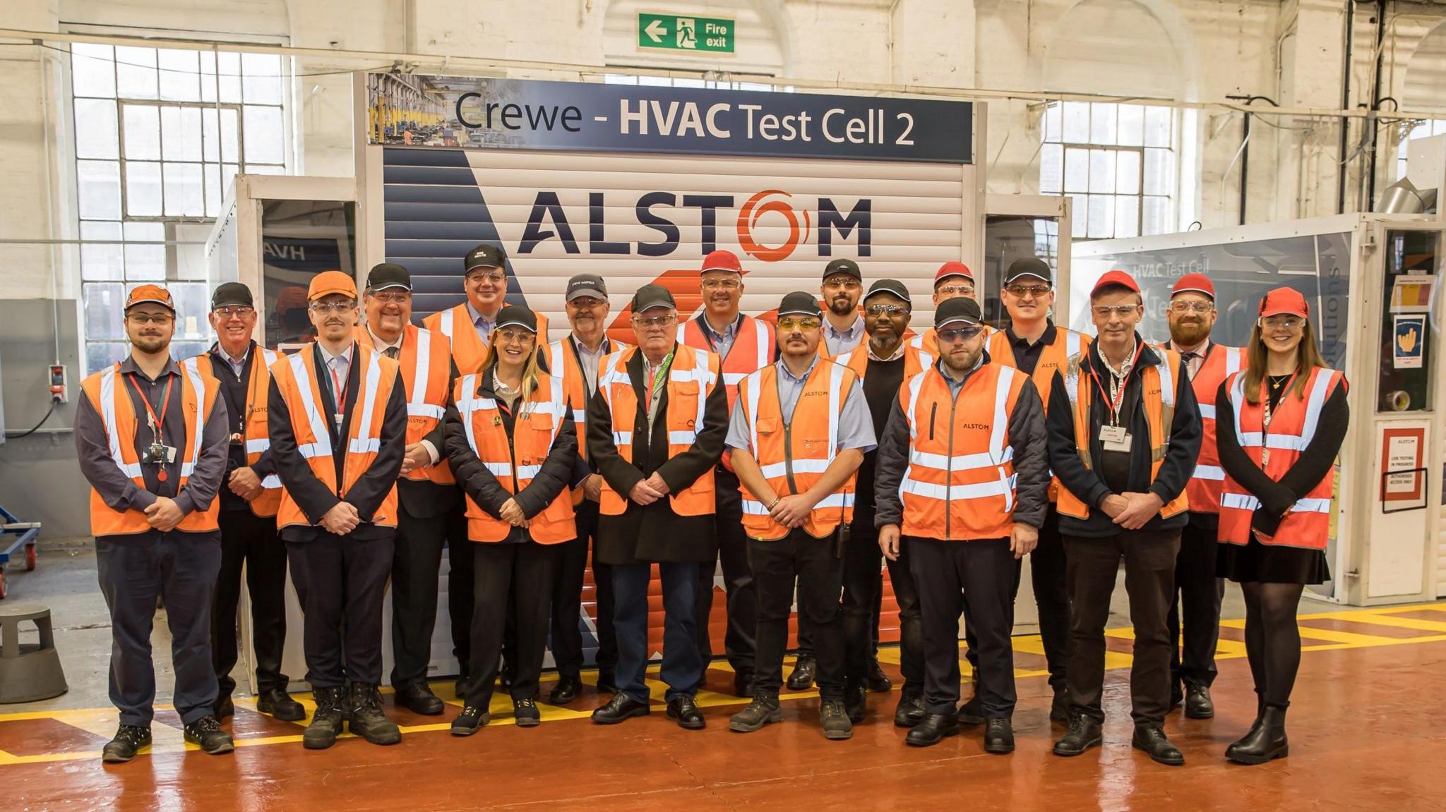 A group of people wearing orange high visibility jackets is standing inside a factory with a polished red floor. There are painted metal shutters behidn them with the Alstom logo on.