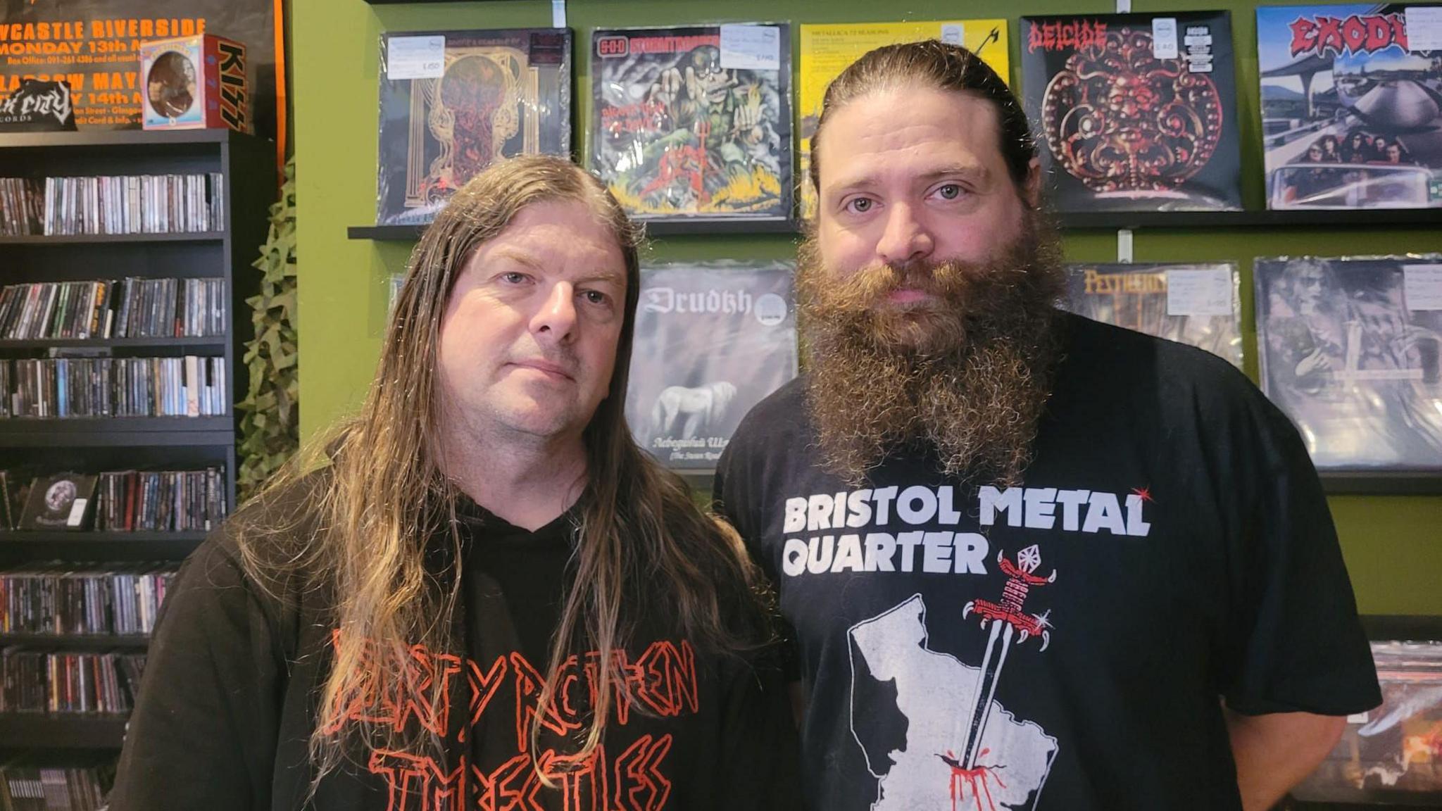 David Savage and John Ashby are standing next to each other in front of a shelf with records. They both have long hair and are wearing black shirts.