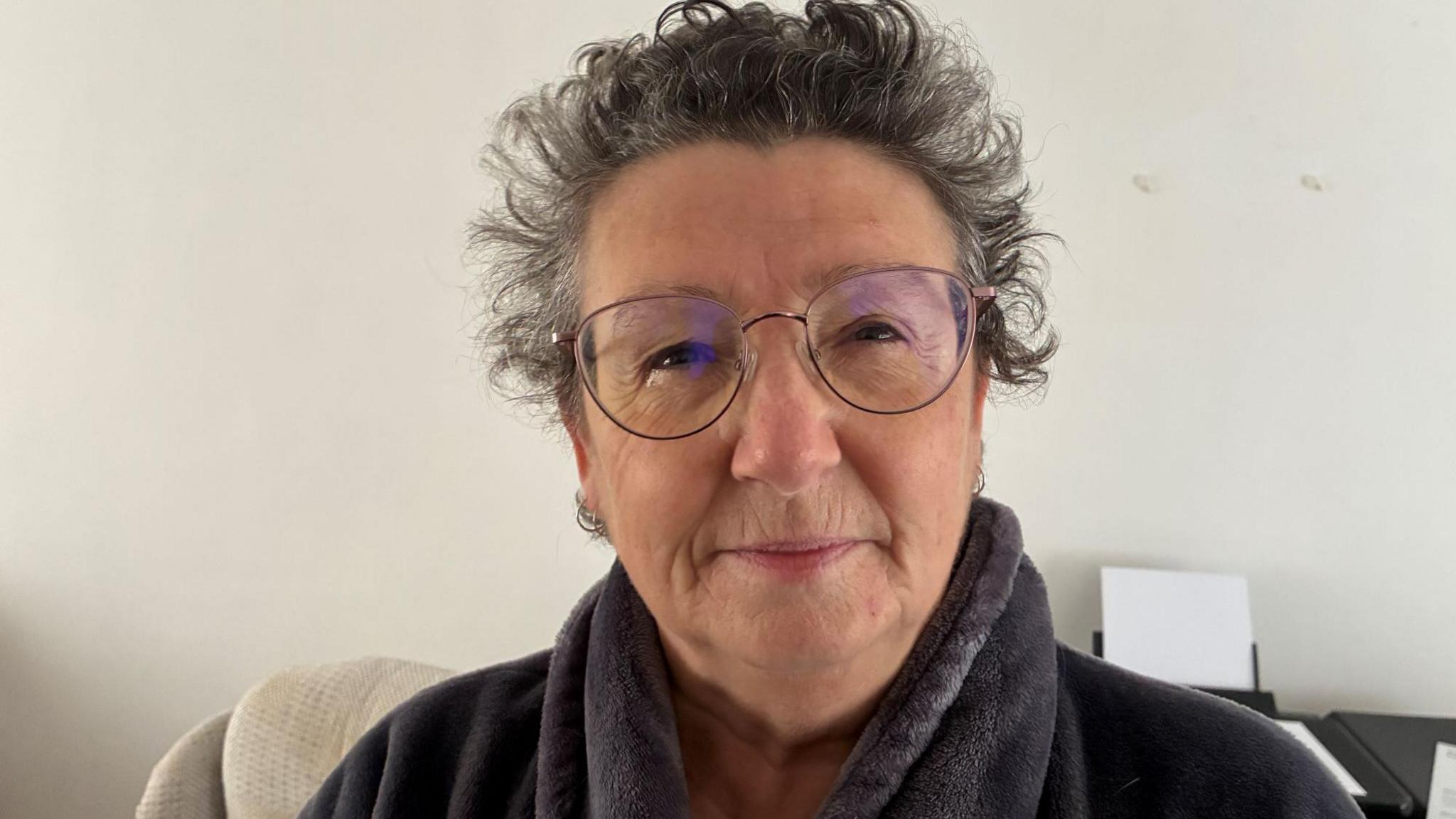 Linda Pendergrass is wearing glasses and a dark grey fleece. She is looking directly at the camera and is in front of a white wall.