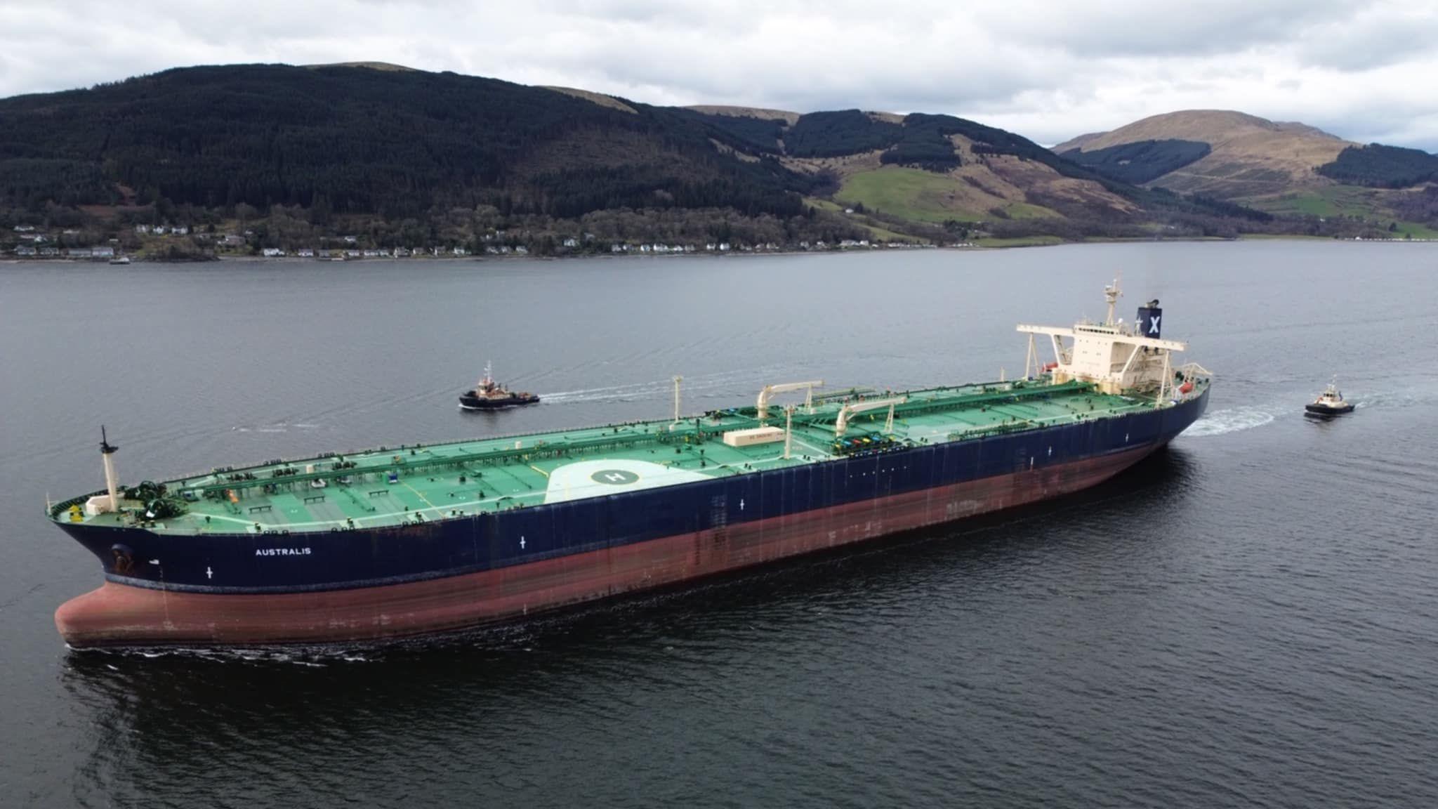 A large oil tanker sails away from the camera. The ship is black and red with green painted decks. Two tugs can be seen behind and to the rear of the tanker