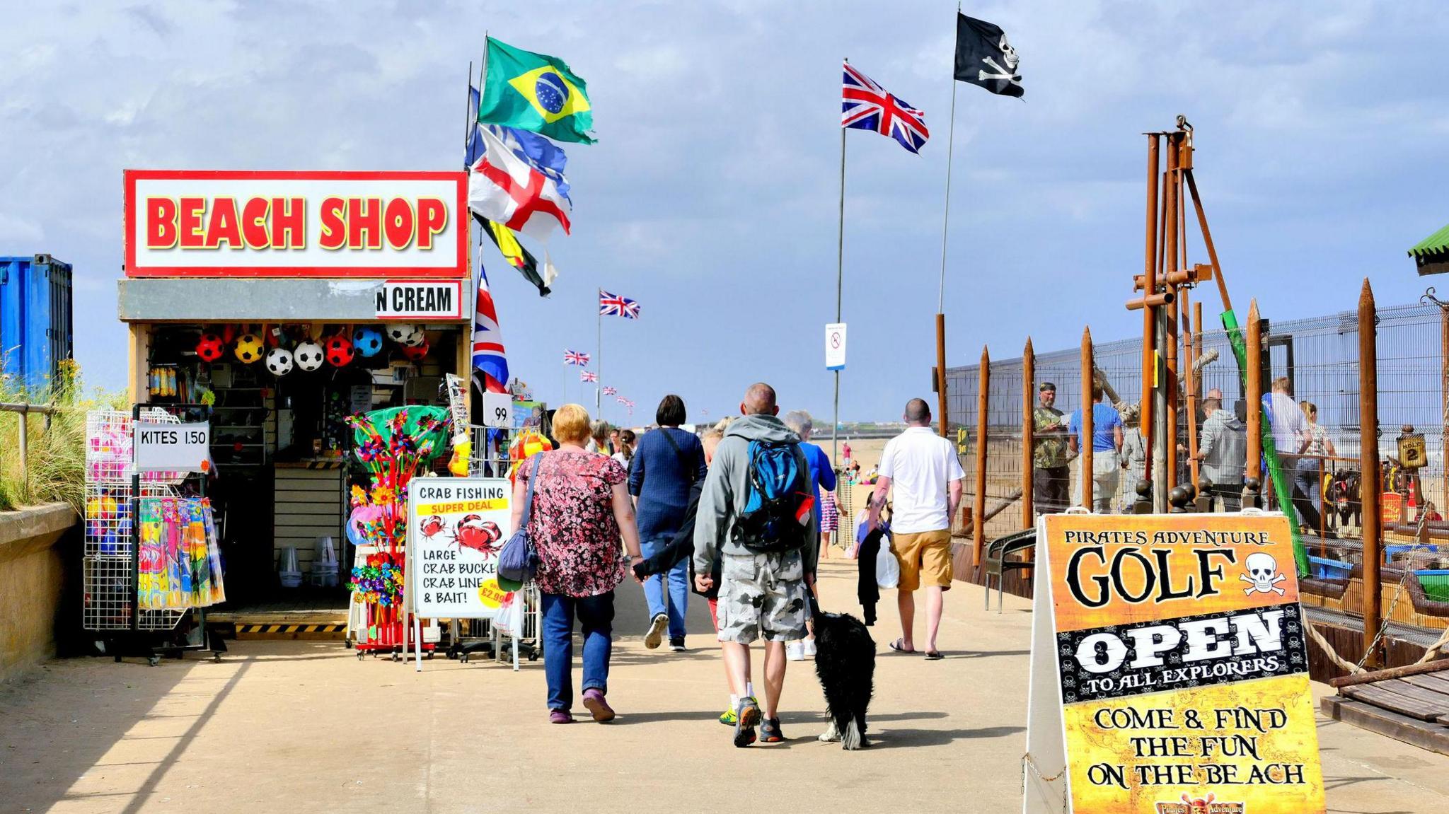 Holidaymakers walking past a beach shop on the promenade at Ingoldmells. There is a billboard advertising adventure golf, along with multiple flags fluttering in the breeze. It is a sunny day.

