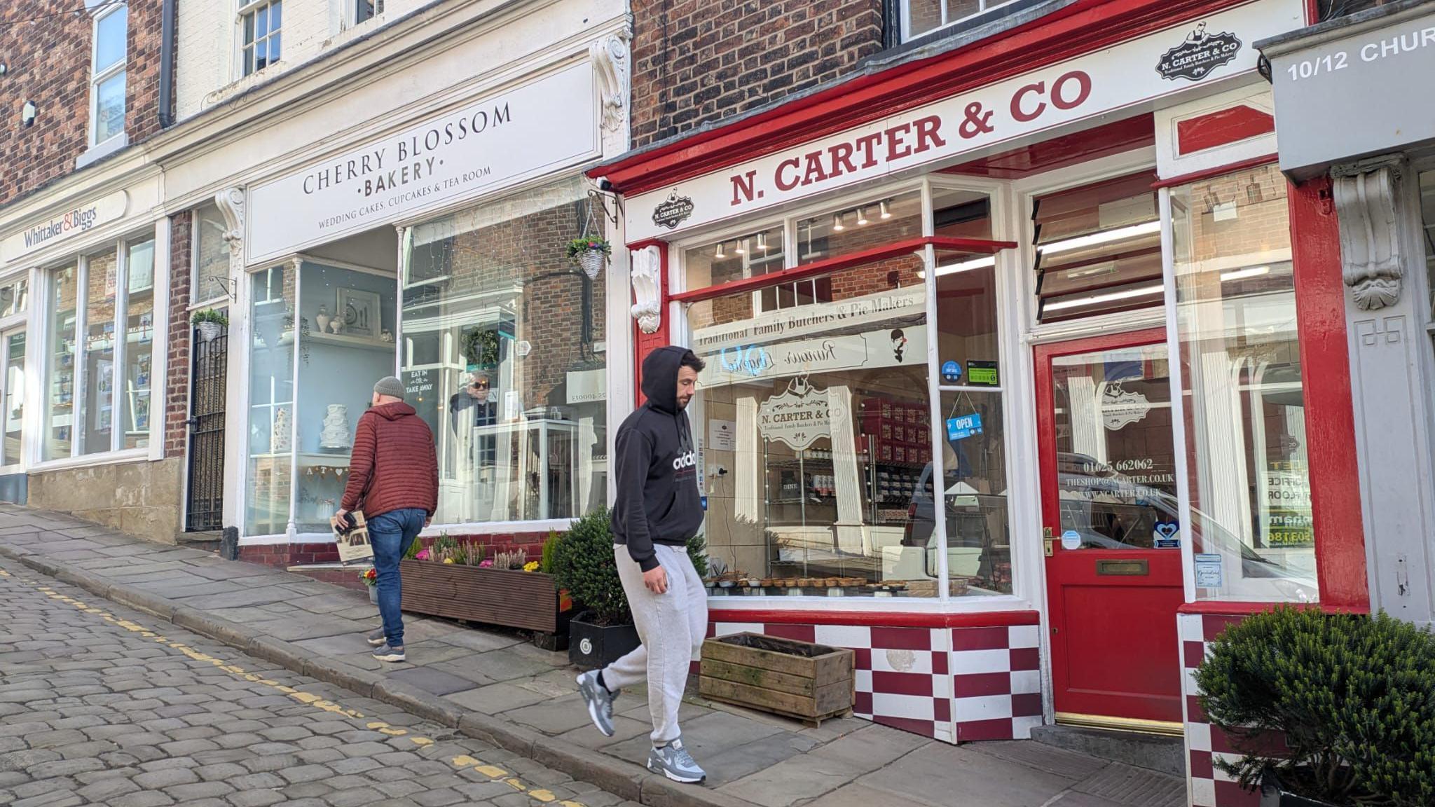 N Carter & Co, a red and white independent butcher alongside a cake shop on a sloping cobbled street in Macclesfield town centre