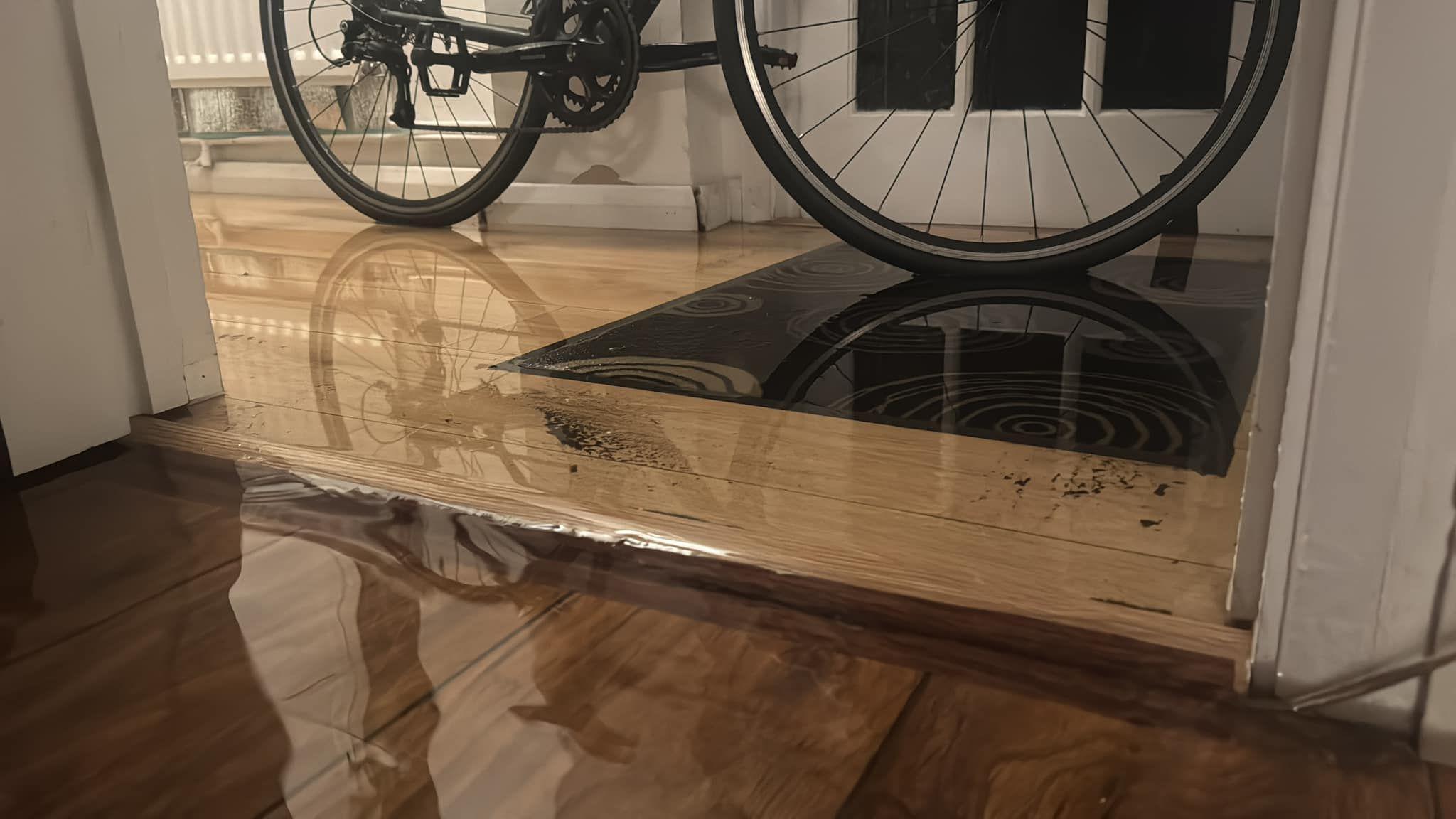 The inside of the house, focussed on the floors. A laminate floored hallway is under an inch or so of water, which reflects the wheels of a black bike that is parked in front of a white and glass door. Older wooden floorboards in the foreground are also under wayer.