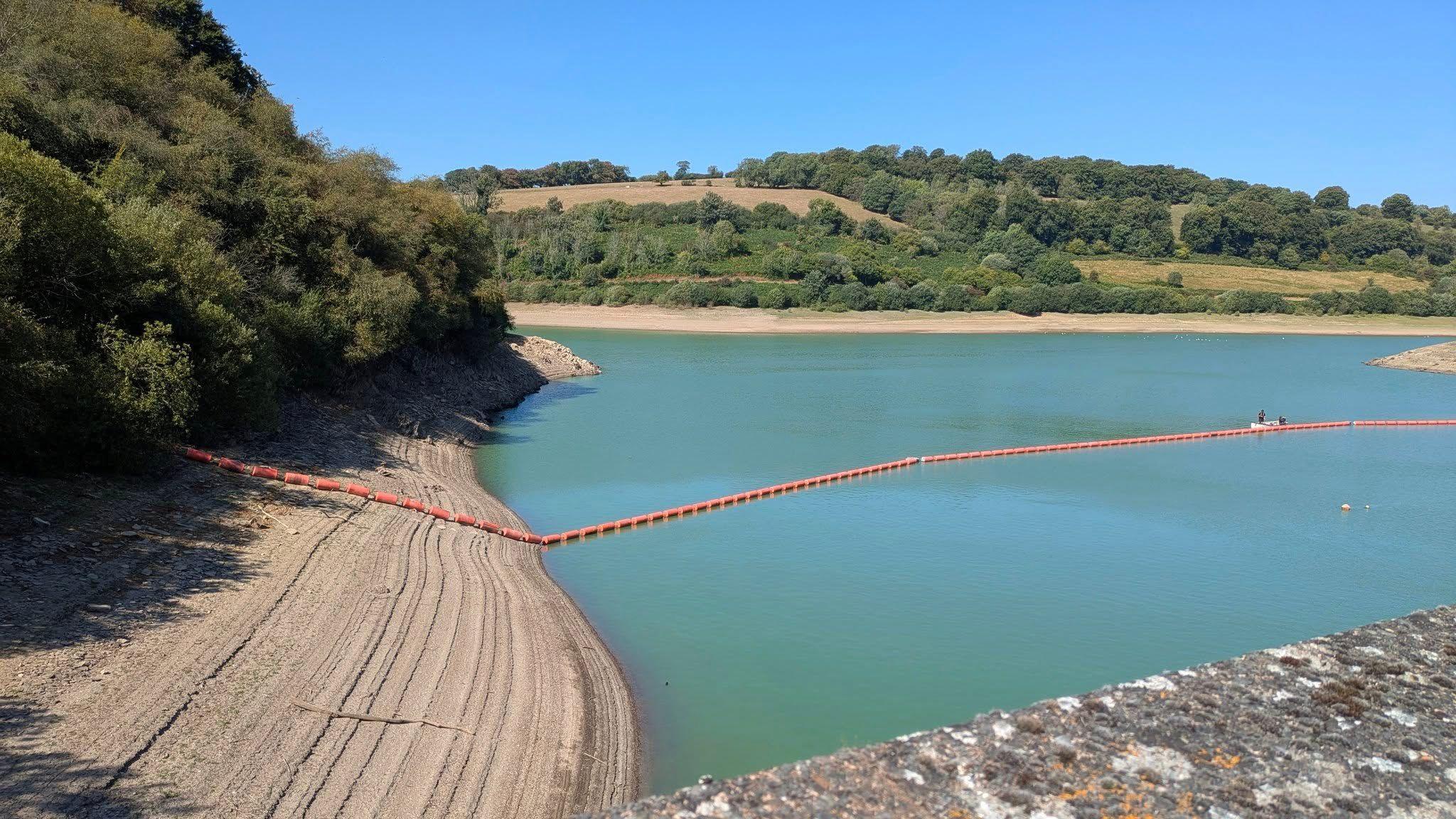 A picture of the low water levels at Clatworthy Reservoir on a bright sunny day with blue skies. There is a strip of exposed bank around the perimeter of the bright cyan coloured water. There is a line of orange floats laying on the sloped dirt bank and stretching across the surface of the water, showing where the water levels usually are. The lake is surrounded by trees and fields which have been scorched in the dry weather. 