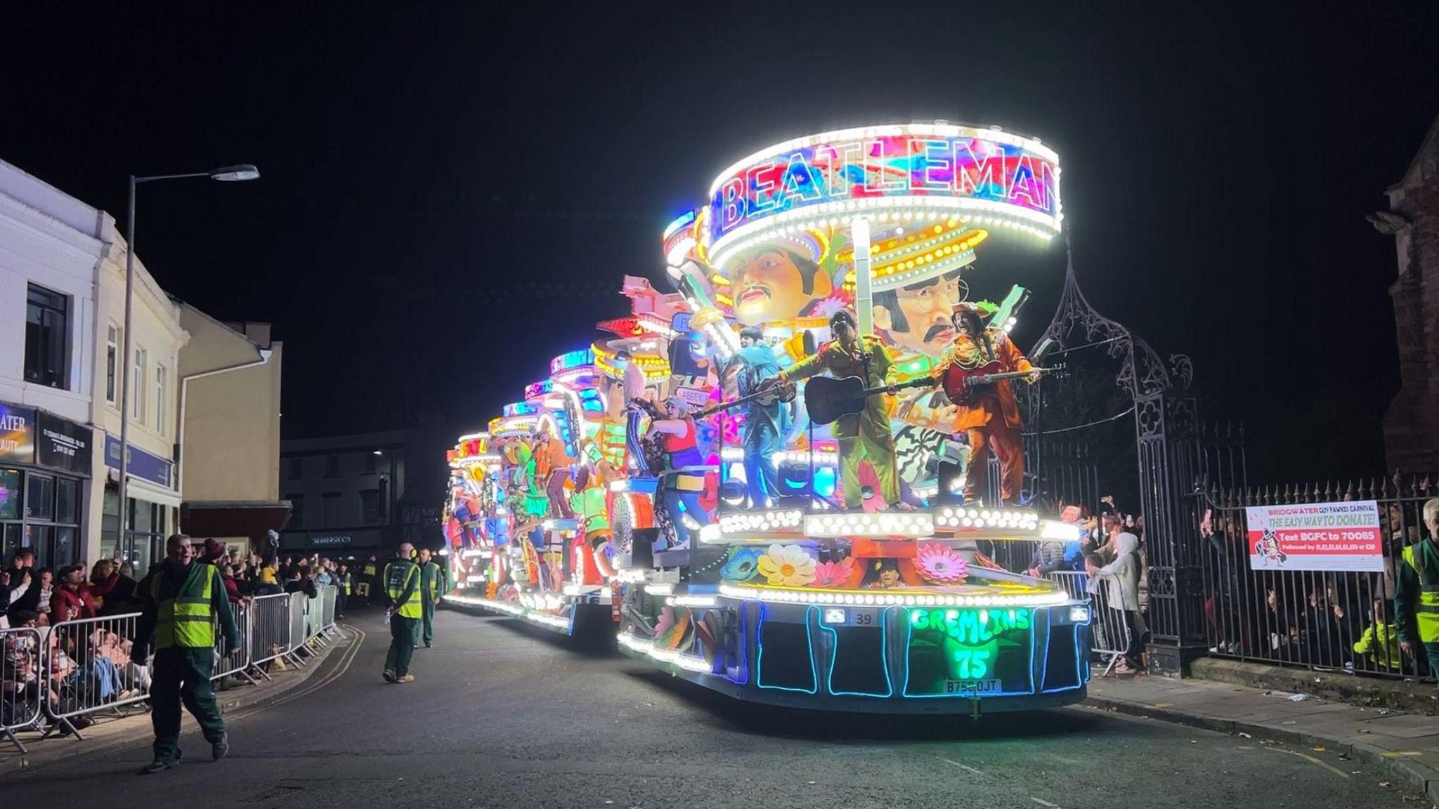 A brightly-coloured and lit up Beatlemania-themed cart coming down the street, with people dressed as the Beatles holding guitars at the front.