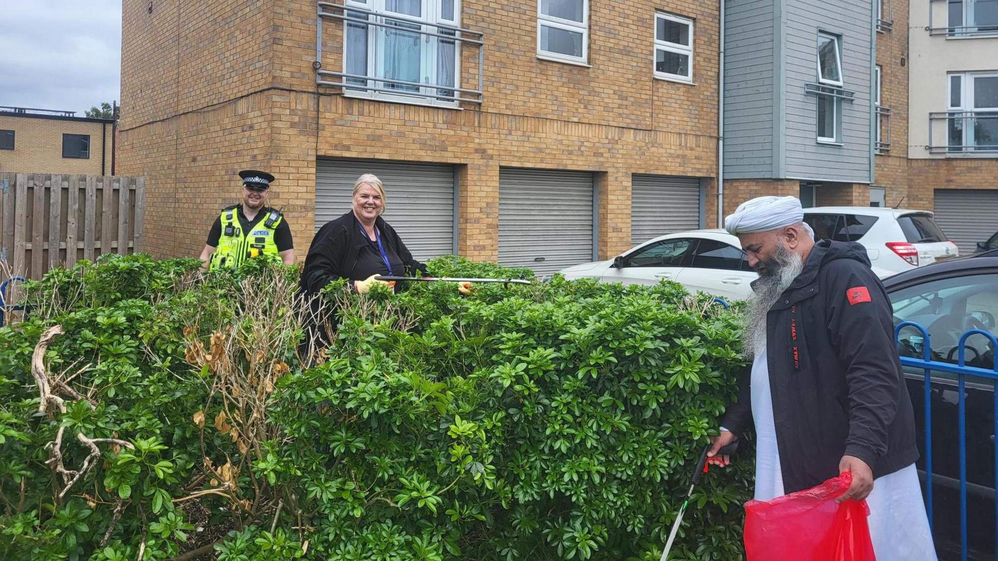 Two people and police officer picking up litter around some bushes