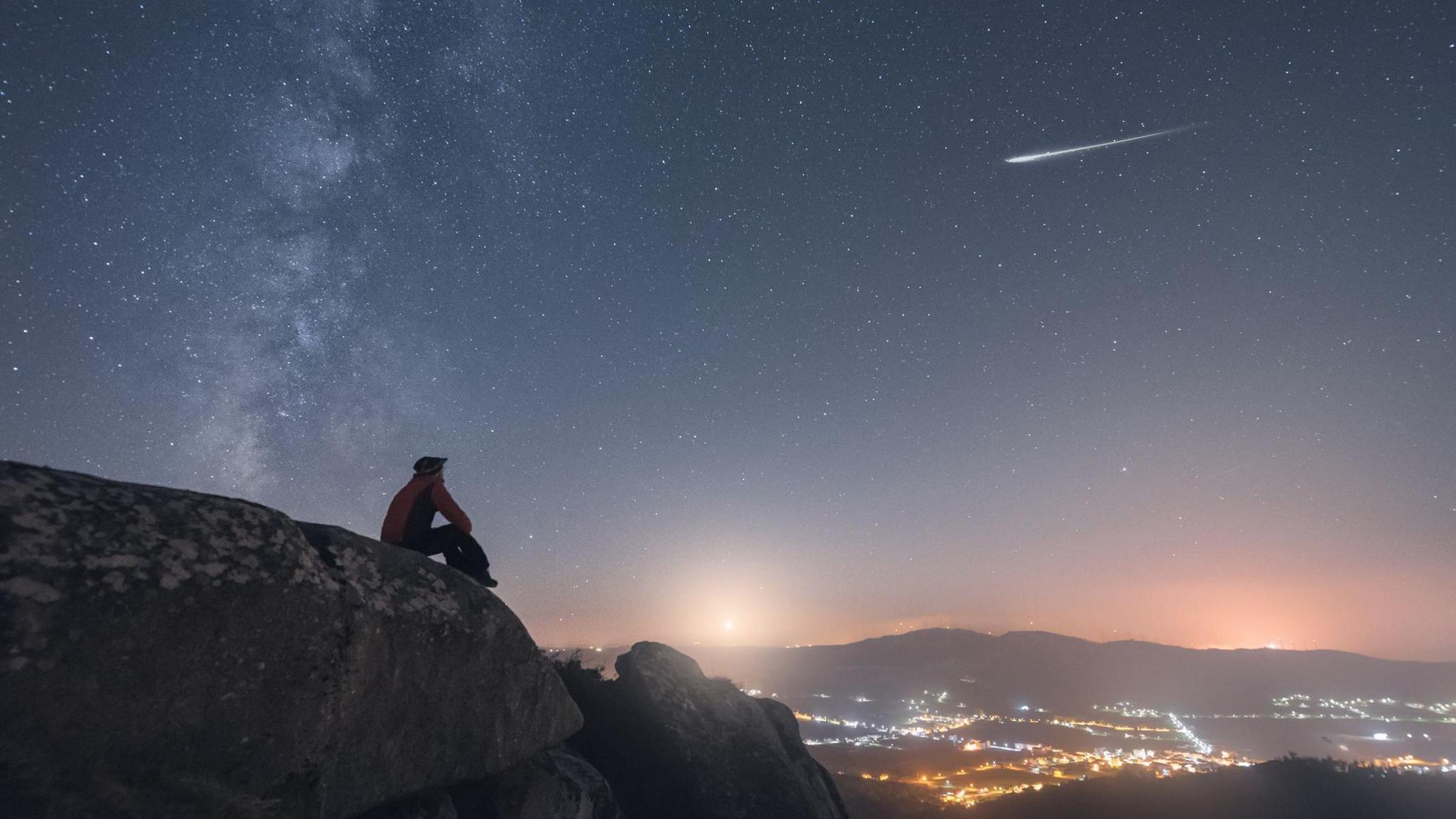 A man sits on a rock above a city with lights below him, watching a shooting star, the milky way and the moonset in the night sky.