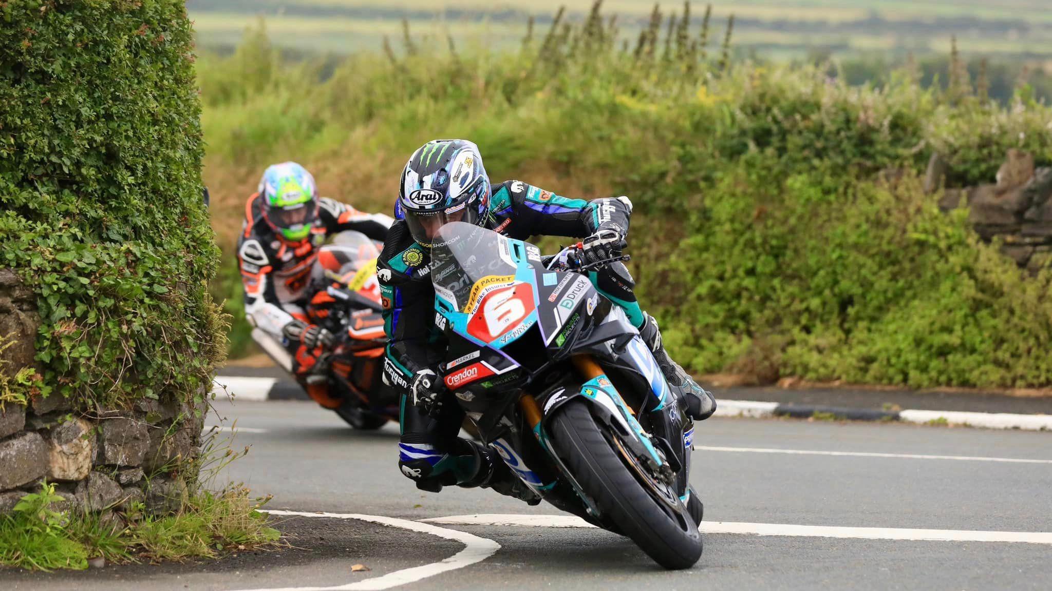 Michael Dunlop racing on the Billown Course. He's leading over on the machine, which matches his leathers and helmet in black, white turquoise and purple with the number six in white on a red background on the front. There is a stone wall covered in green foliage on the left and a sod hedge covered in grass behind him. He is being followed on the road by Jamie Coward, who is also on a bike that matches his leathers in white, orange and black. His helmet is green and yellow.