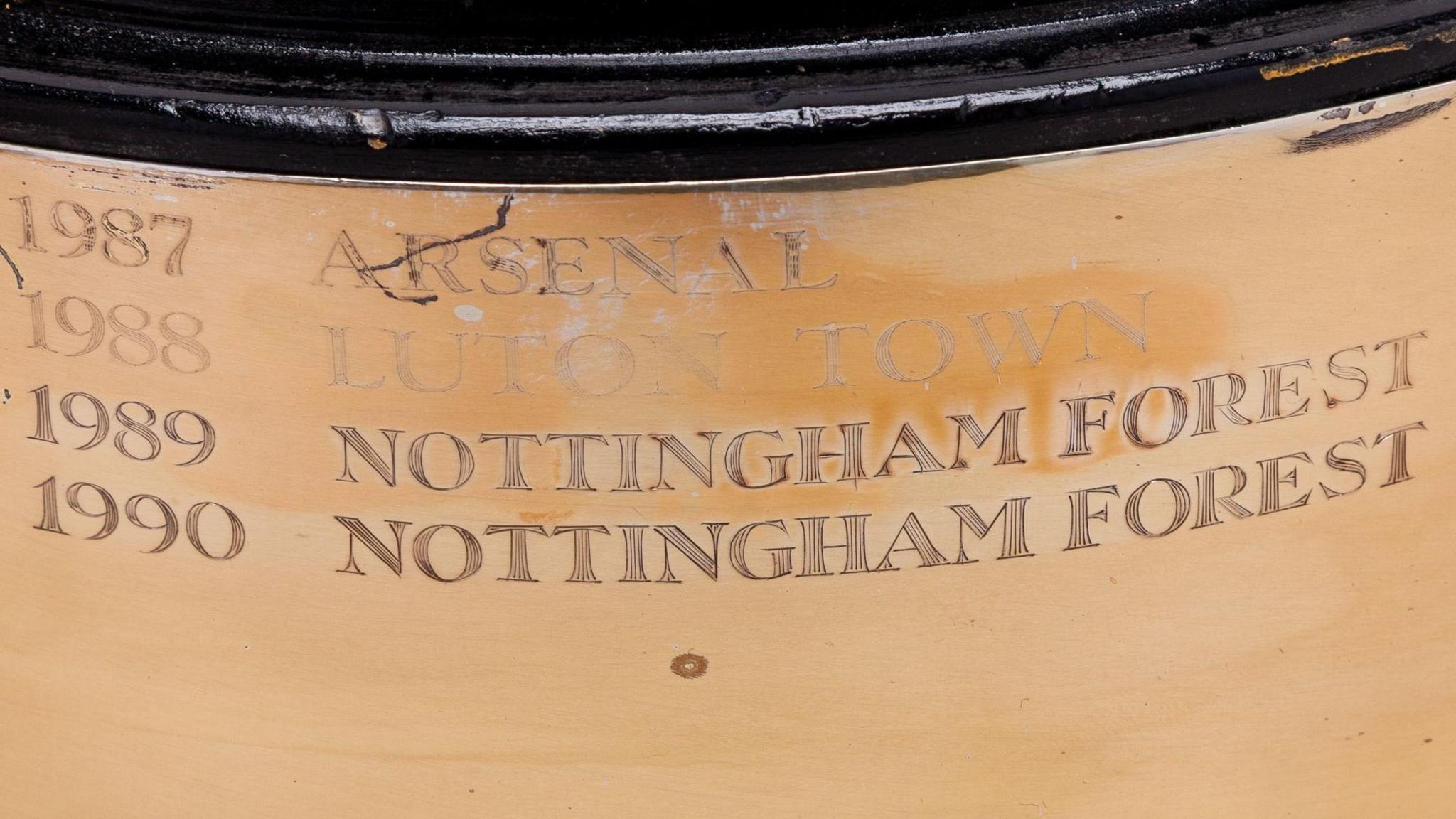 A close-up of the four names of the three clubs to win the trophy, etched on to its gilded base with their dates. The dates are on the left and the names on the right in capitals. From the top 1987 Arsenel, 1988 Luton Town, 1989 Nottingham Forest, 1990 Nottingham Forest.