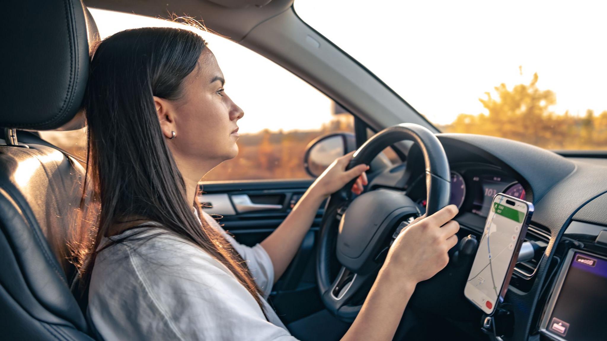 A young woman with long, straight, dark hair driving a car and using a hands-free navigator on her smartphone (stock image) 