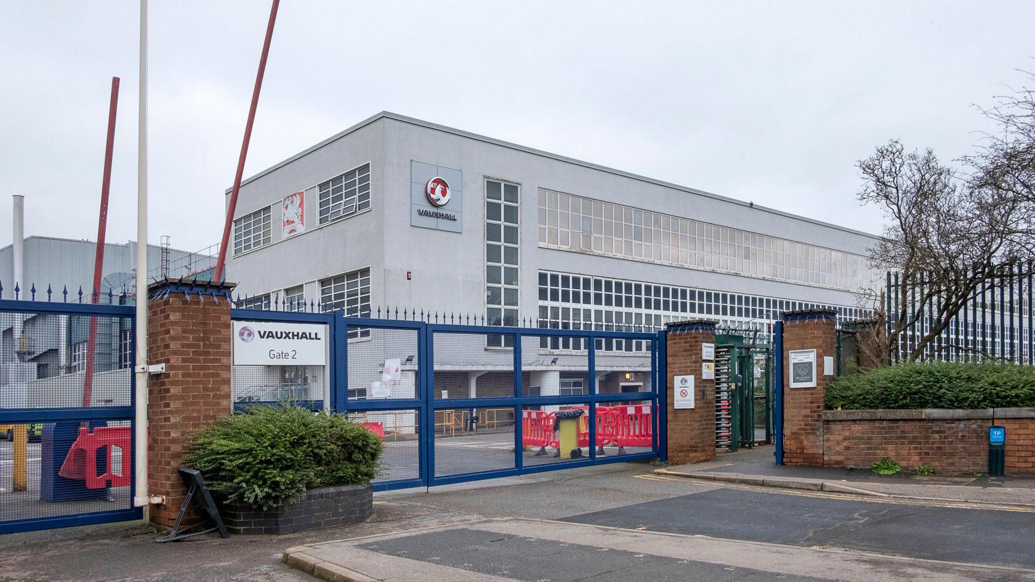 The entrance to the assembly plant of the automotive manufacturing company IBC Vehicles Ltd, operating under Vauxhall Luton and producing light commercial vehicles, in December 2021. It shows blue metal gates, red barriers, and a grey building with lots of windows 