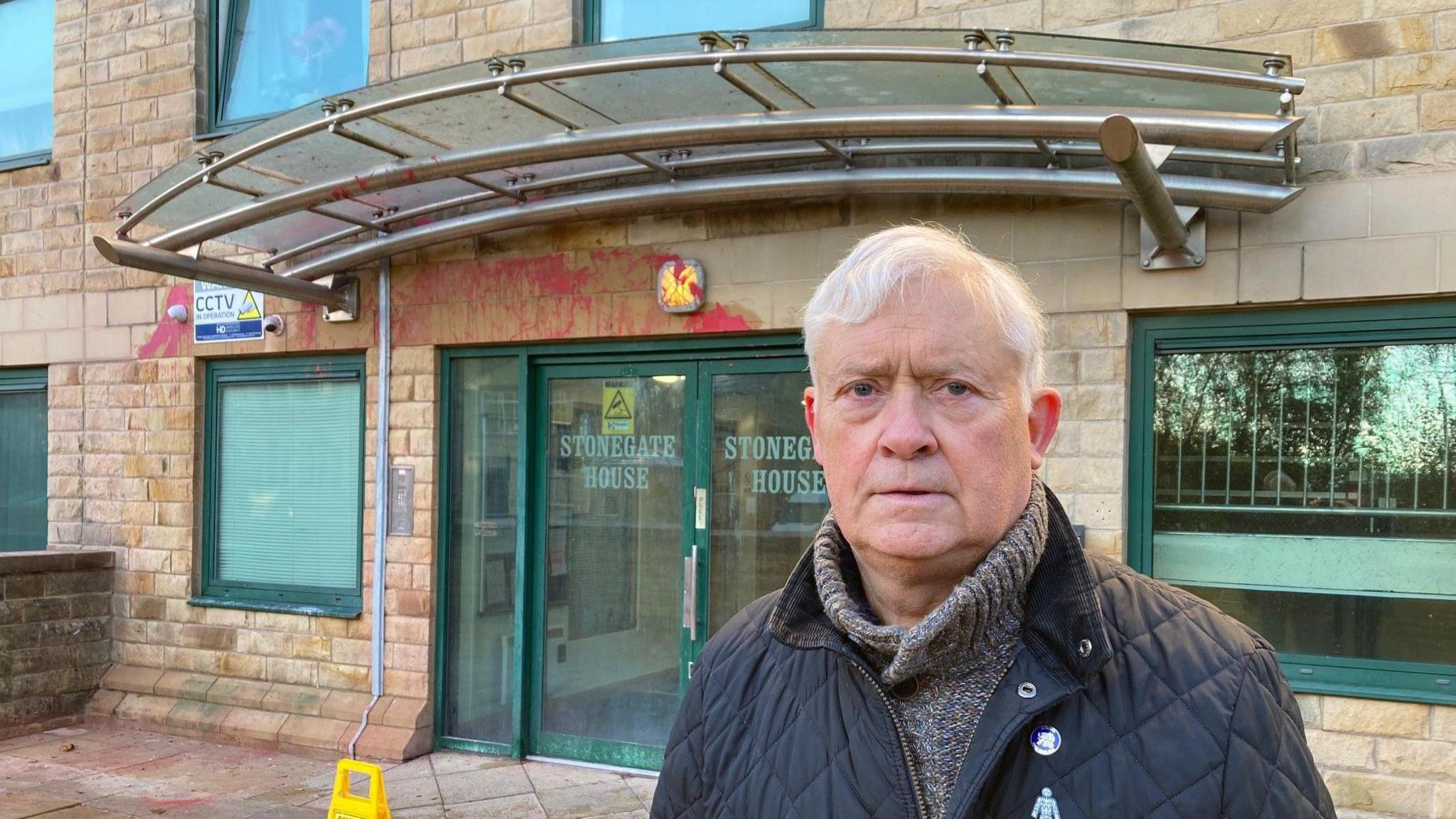 A white-haired man wearing a blue quilted jacket and grey jumper underneath outside an apartment block entrance with green-framed doors and glass panels with the remnants of red paint above the doors. 