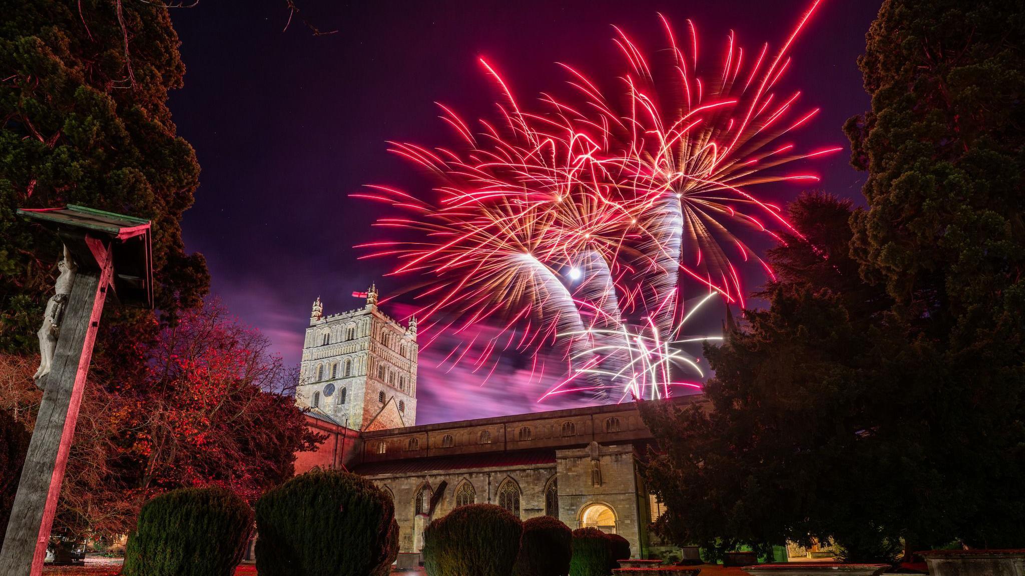 Pink, white and purple fireworks light up the dark sky behind Tewkesbury Abbey which is also lit up. The image is taken from ground level.
