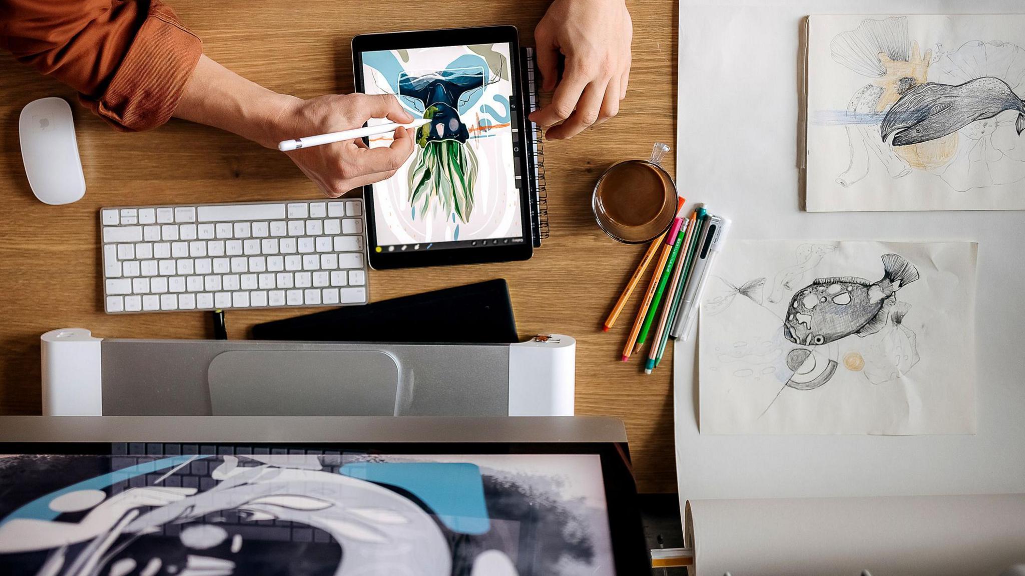 A person is drawing a plant on a black tablet next to a keyboard and mouse. There is a mug on the table and other sketches.