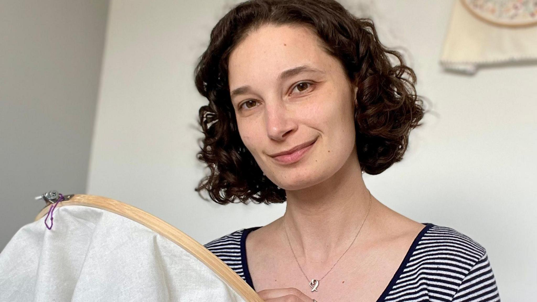 A woman with a short bob and a stripy black and white t-shirt smiling at the camera while holding her embroidery hoop.