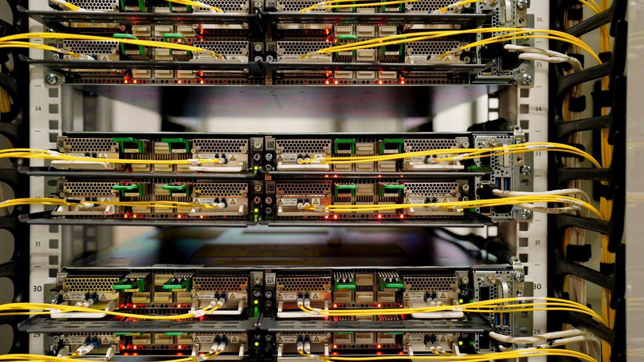 Wiring is plugged into the back of different machines in a stack. The wires are yellow and small red lights are underneath some of the wires.