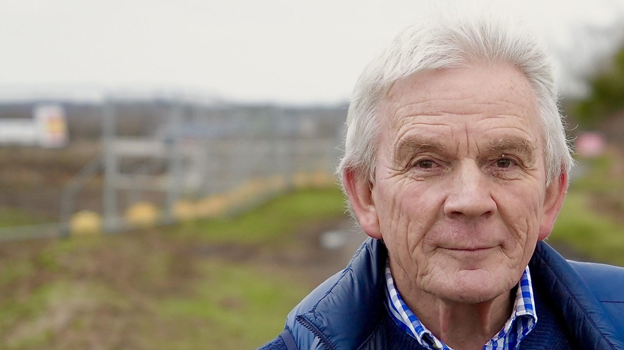 David Grant stands by fields that have been cleared for a Sizewell C access road. He has grey hair and is wearing a blue gilet over a blue sweater and a blue and white checked shirt.