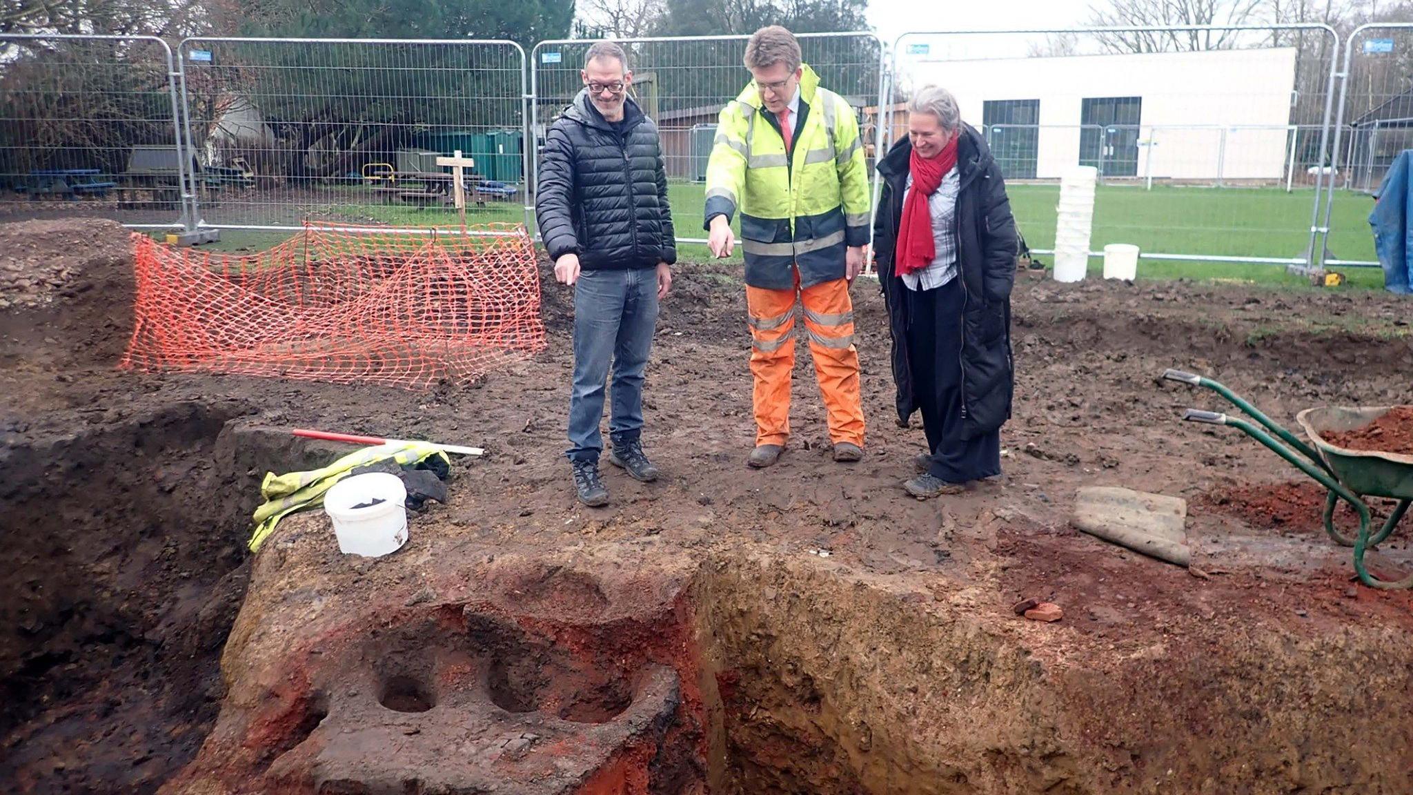 Two men, one dressed in black and another wearing high-vis protection on top of his suit, are pointing at the dig site. A woman wearing a long black coat and a red scarf is stood next to them