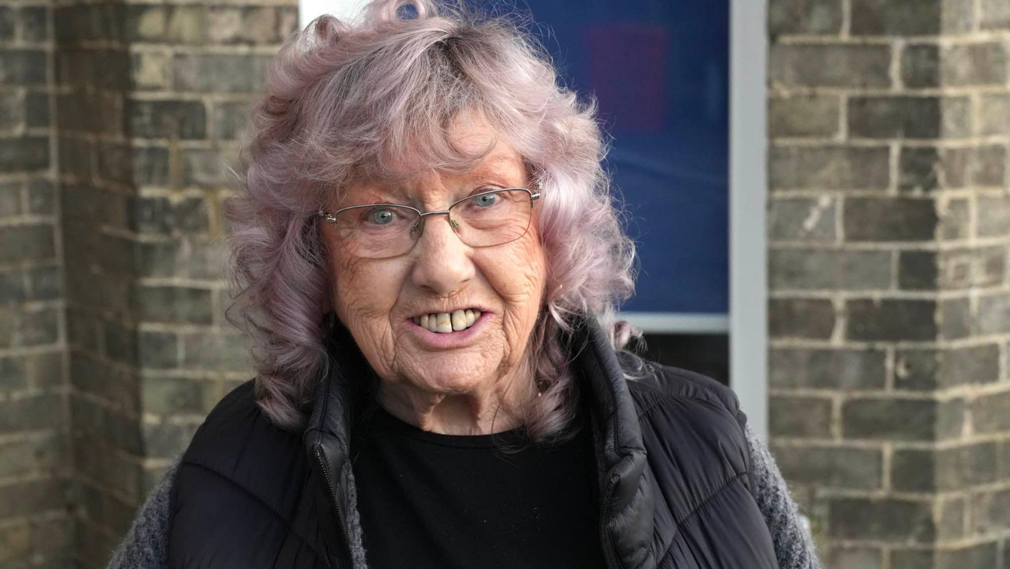 A woman with shoulder-length light purple hair and glasses. She is smiling into the camera.