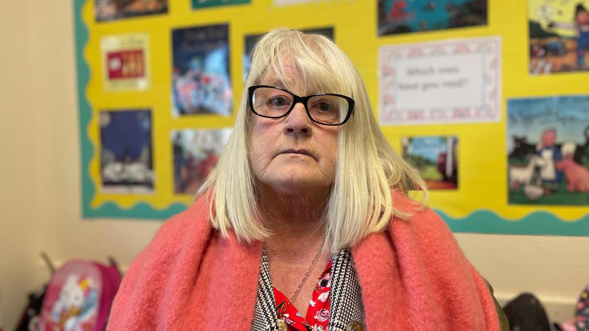 A woman with fair shoulder-length hair and dark-framed glasses. She is wearing a pink shawl over a black and white jacket. Behind her on the wall is a yellow board with a green border and images of covers of children's books.