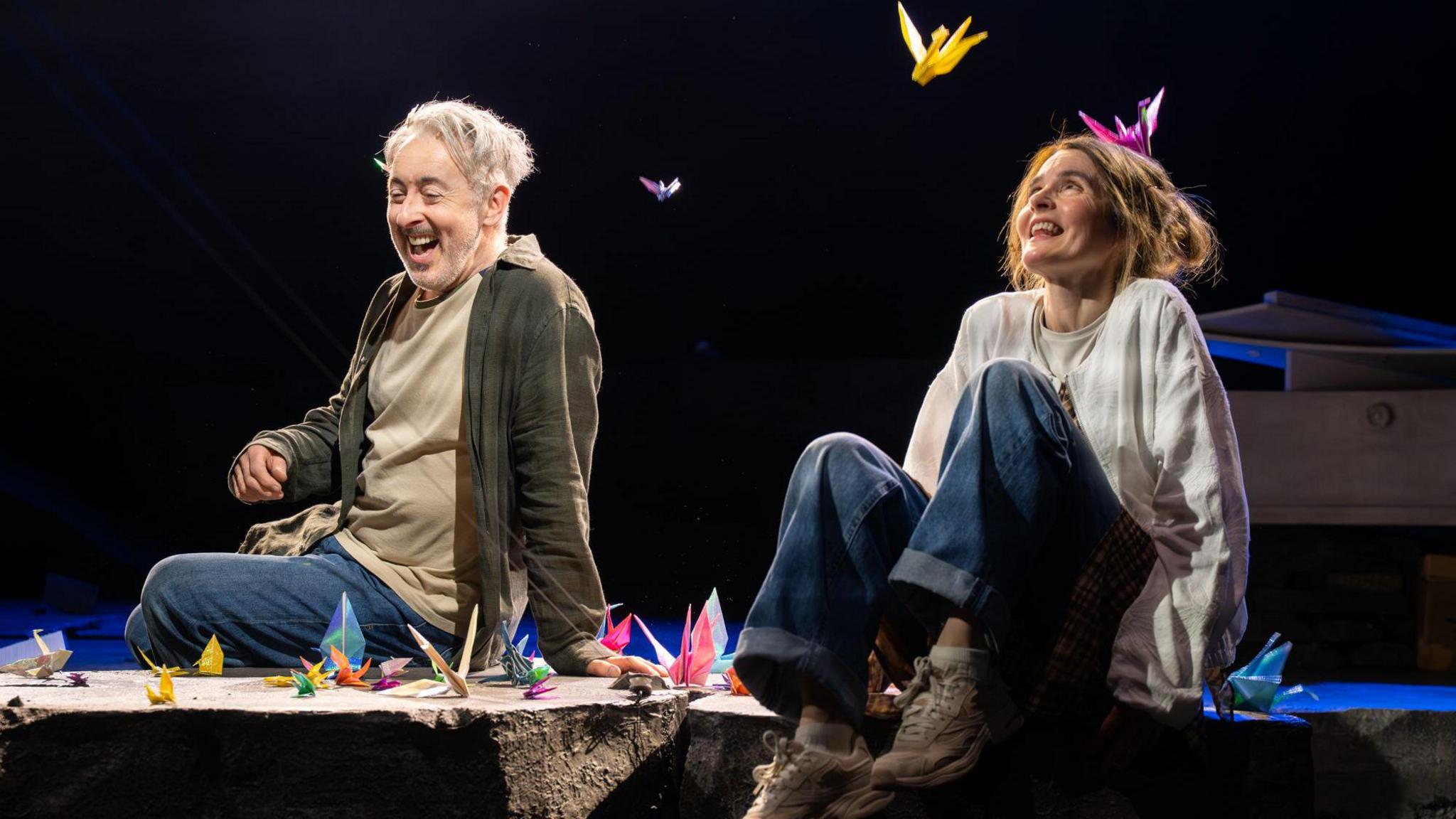 Two performers sit on the edge of a stage set, surrounded by brightly coloured origami cranes scattered around and suspended in the air. Dramatic spotlights illuminate the scene against a dark background, drawing attention to the vivid paper sculptures and casual costumes.
The theatrical setting creates a striking contrast between the simple staging and the whimsical, dreamlike display of floating paper birds.