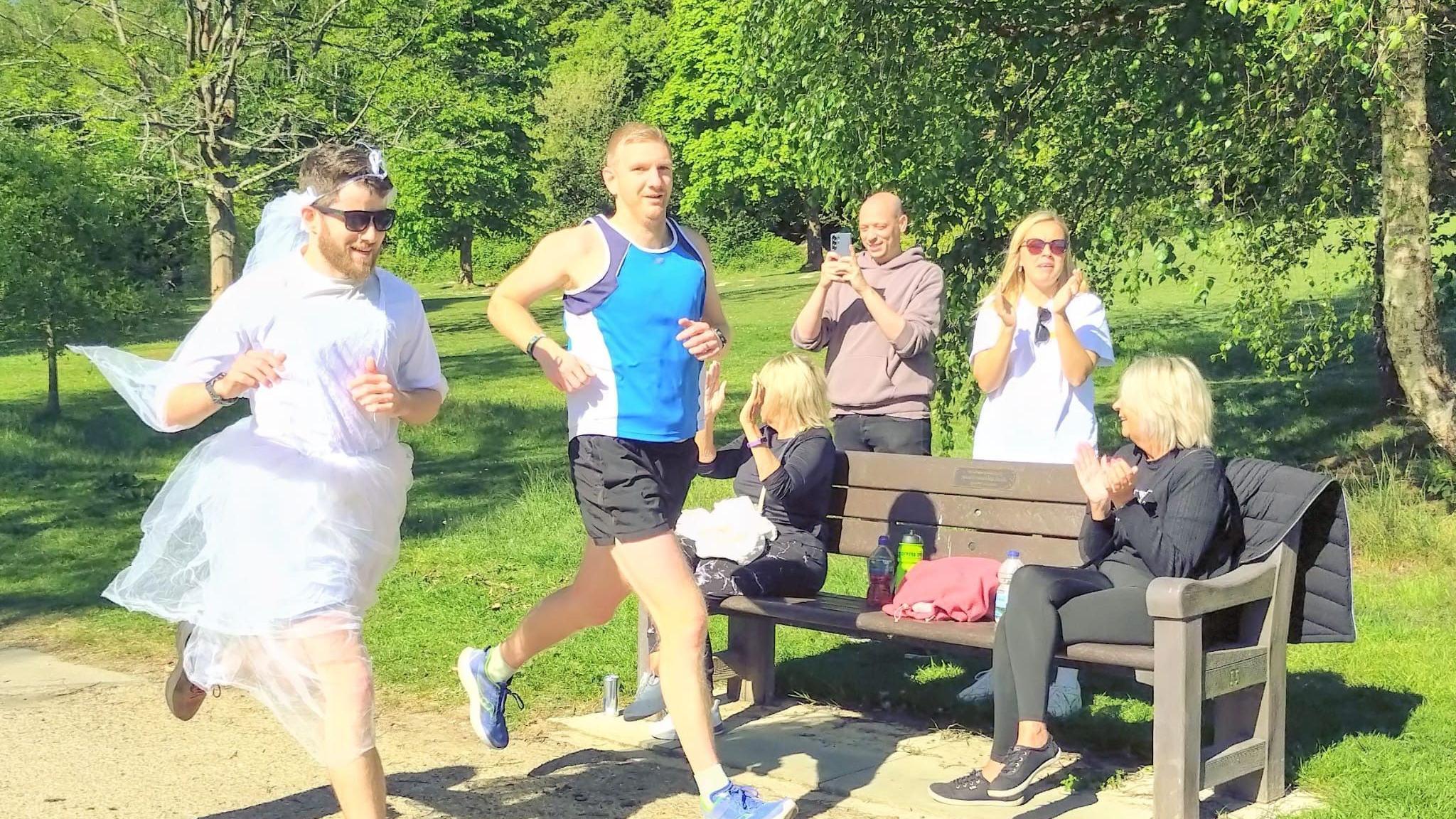 Two men running while onlookers clap and cheers. One man is wearing a white wedding dress and sunglasses while the other is wearing a blue top and black shorts.
