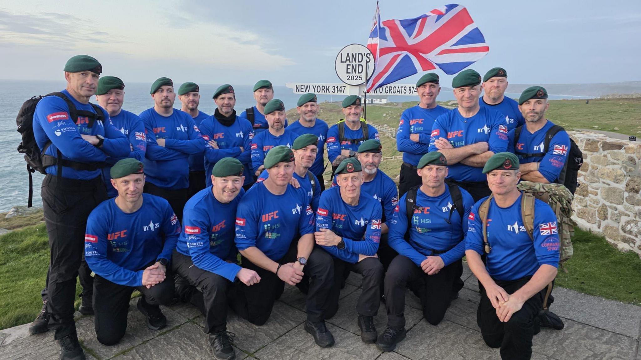 A group of veterans stand and kneel at Land's End. The group are looking at the camera. A Union Jack flag is flying behind them.