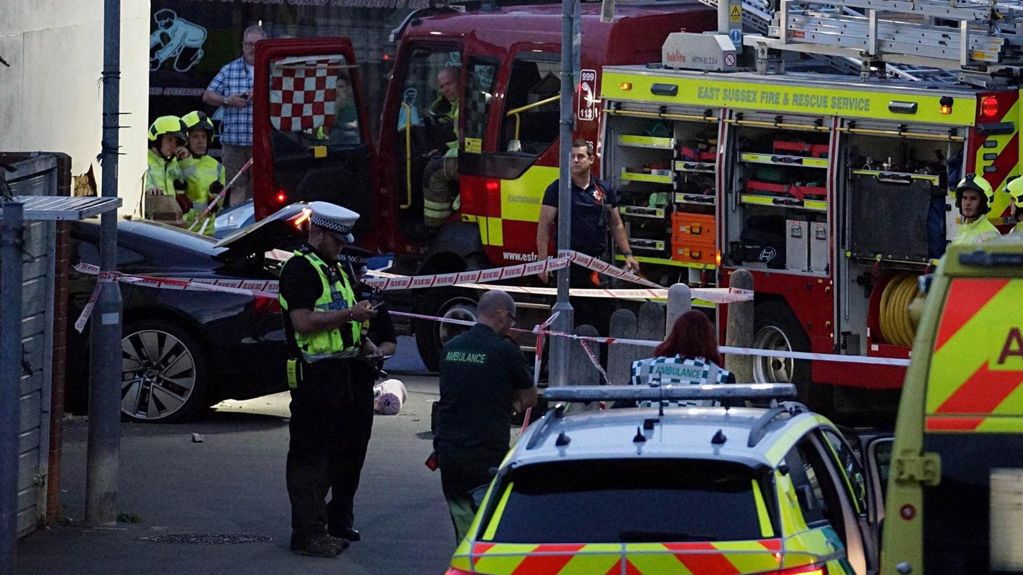 Emergency service crews surround a black car that has crashed into a white wall. 