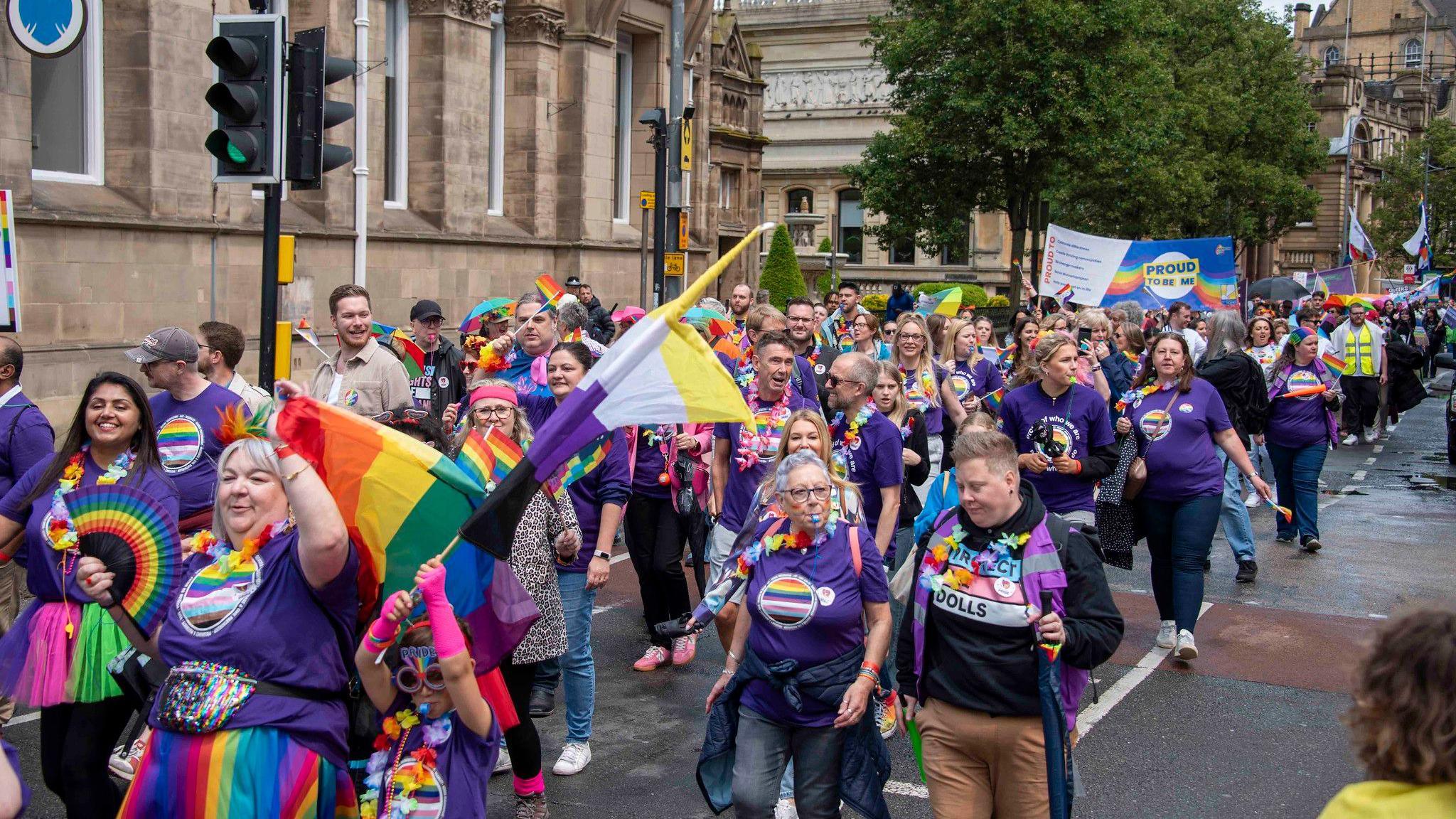 A huge crowd of people parading down a city centre street, Most are wearing purple shirts and rainbow colours. Some are holding banners or waving rainbow-coloured flags.