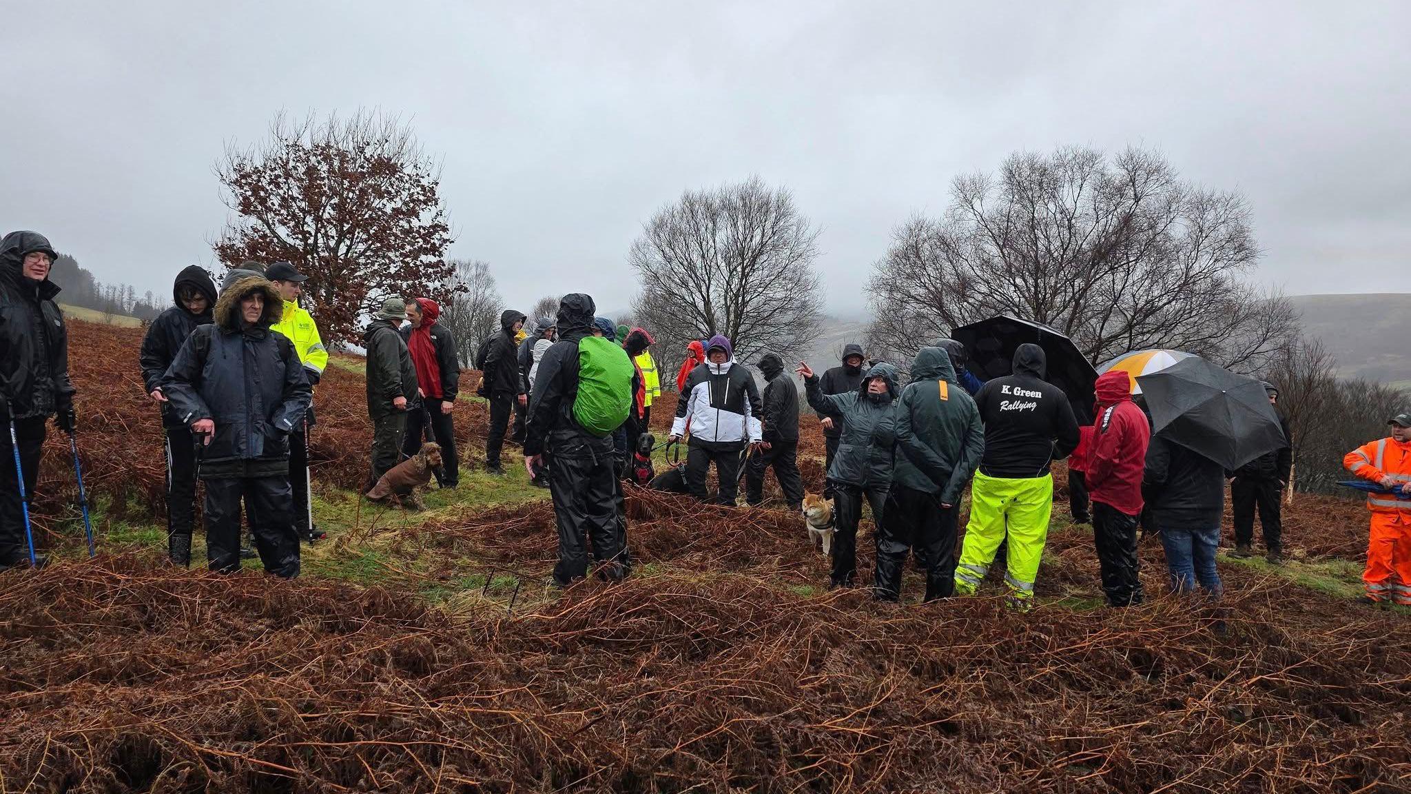 A picture of a group of around 30 people gathered on a mountain side with grey sky surrounding them, appearing sodden with rain. Some have dogs on short leads and others have walking sticks. 
Around three people can be seen holding umbrellas while everyone wears raincoats and has their hood up. 