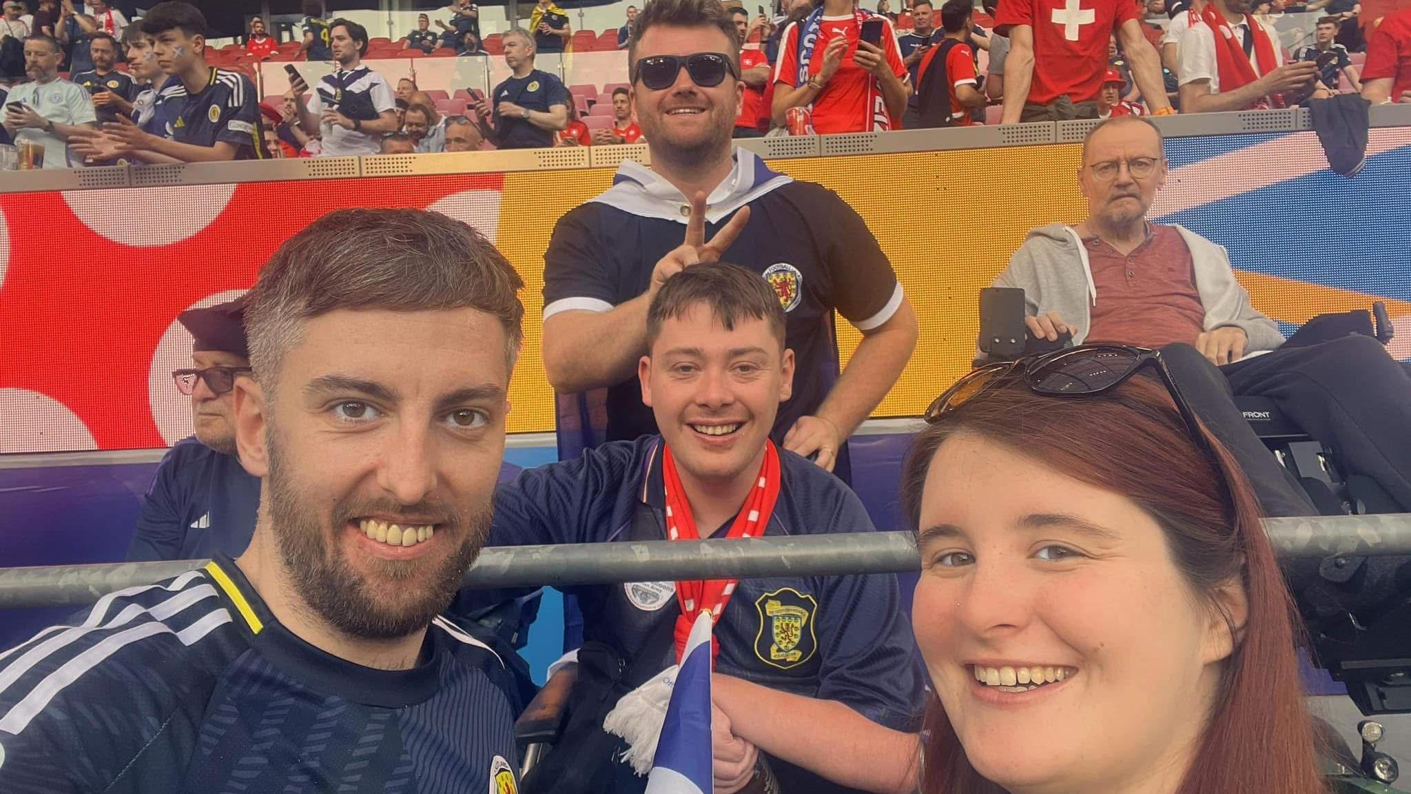A smiling Stephen (centre) sits in the stand for a selfie style pic taken by a fellow supporter. He is wearing a Scotland shirt. A man and woman, both smiling, are in the foreground. A smiling man is standing behind Stephen and making a v-sign behind his head.