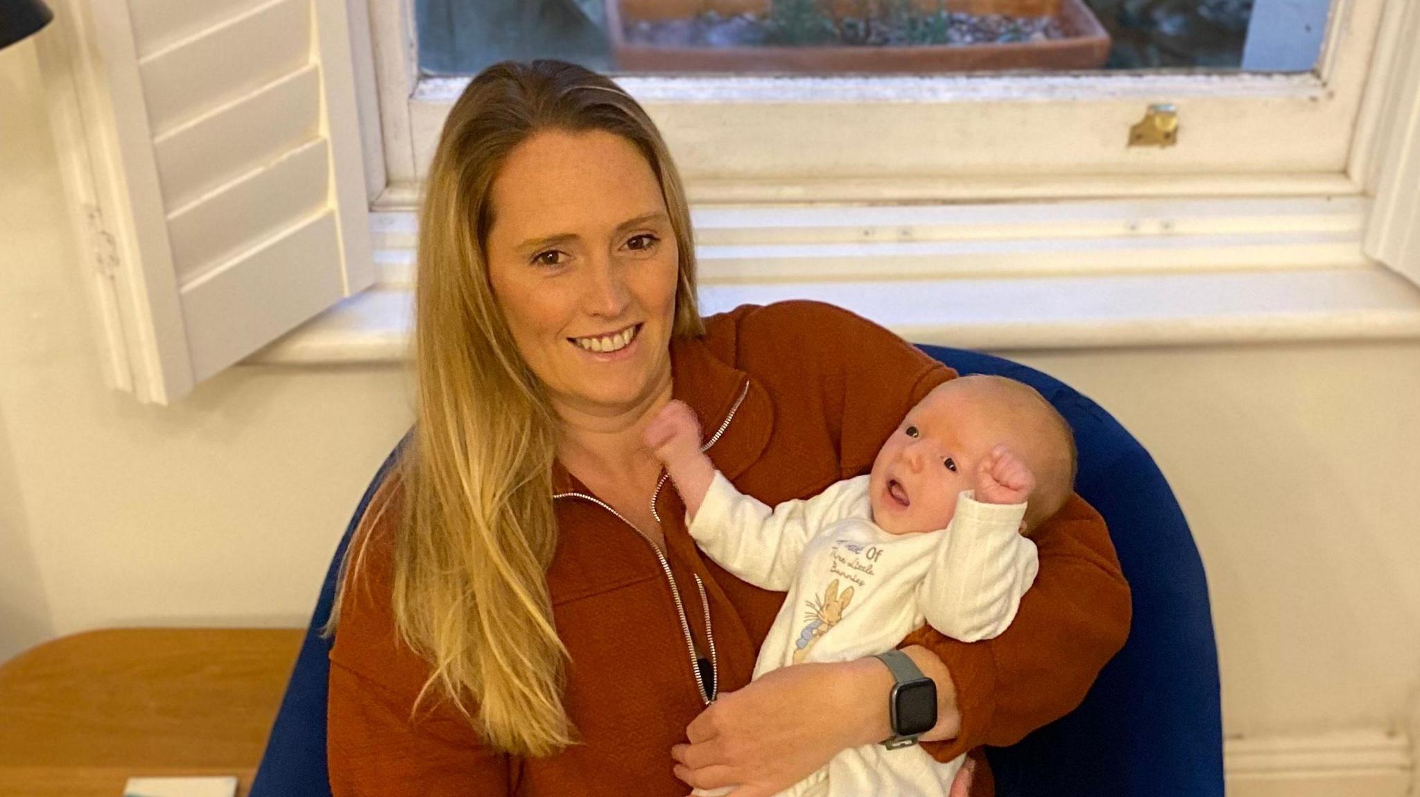 Tamara Vodden wearing a brown cardigan sitting in a blue chair holding her baby, who is wearing a white onesie and holding his arms up. 