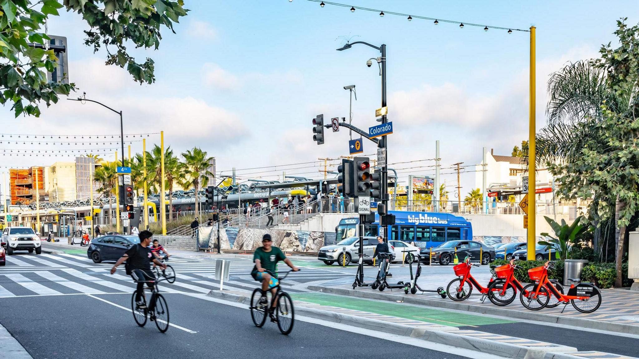 Two cyclists on an LA road ride through a traffic light while rentable red bikes are pictured on the sidewalk