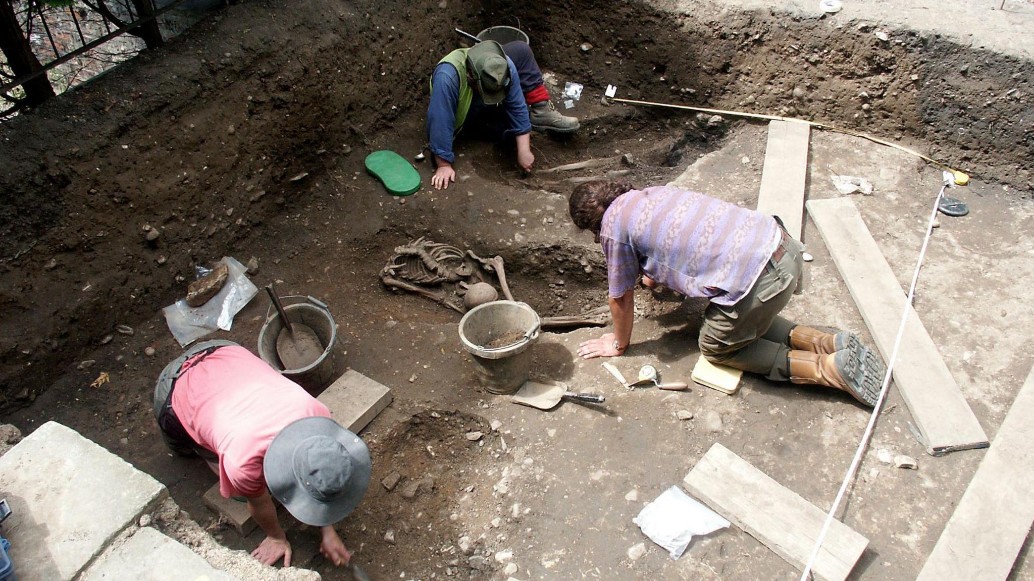 Three men crouched down in the ground during an excavation dig. Skeletal remains are lying in the soil and there are buckets, shovels and planks of wood on the ground.