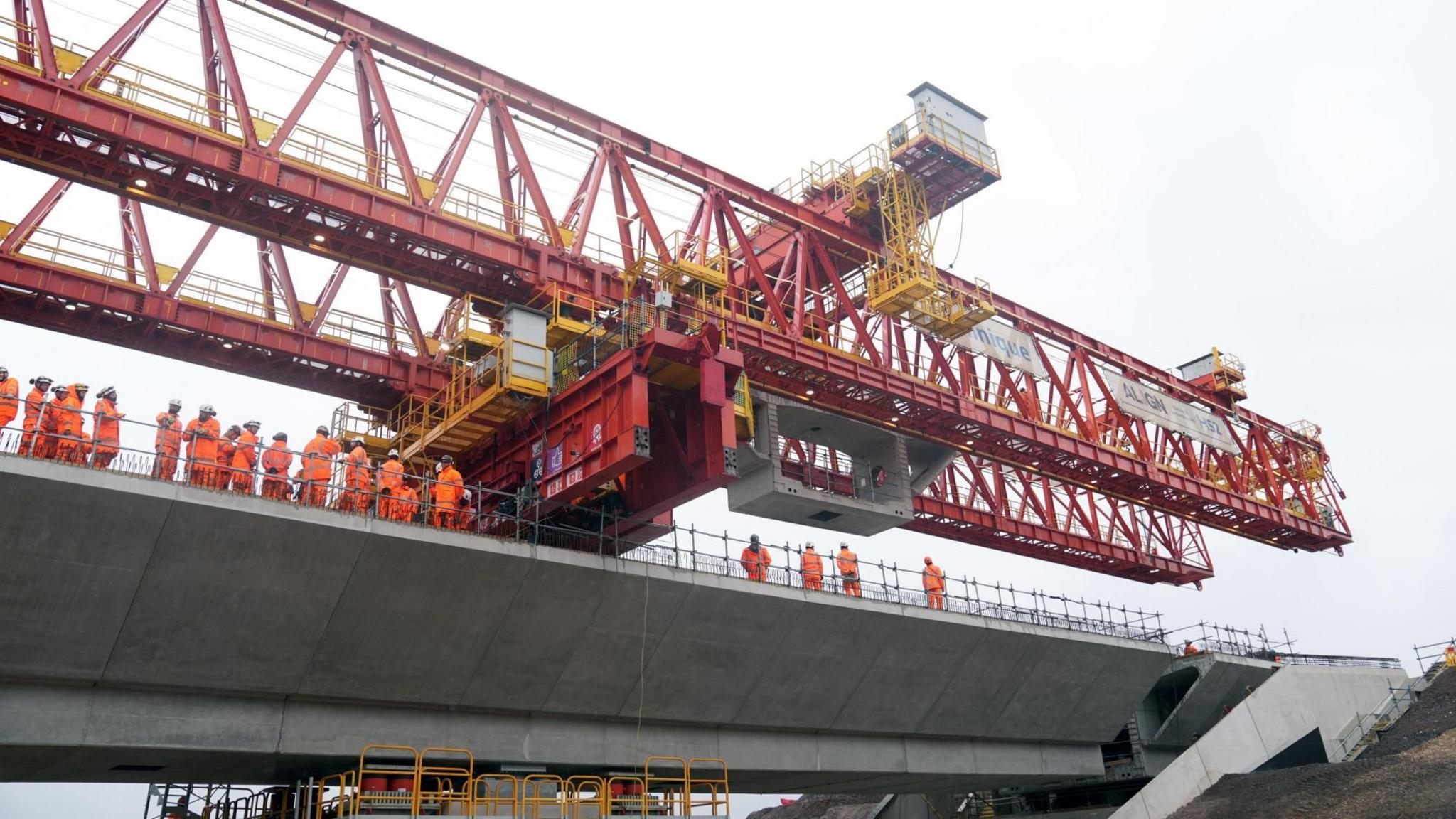 Workers wearing orange high-vis outfits and helmets stand on the grey concrete bridge. Red construction equipment towers over them.