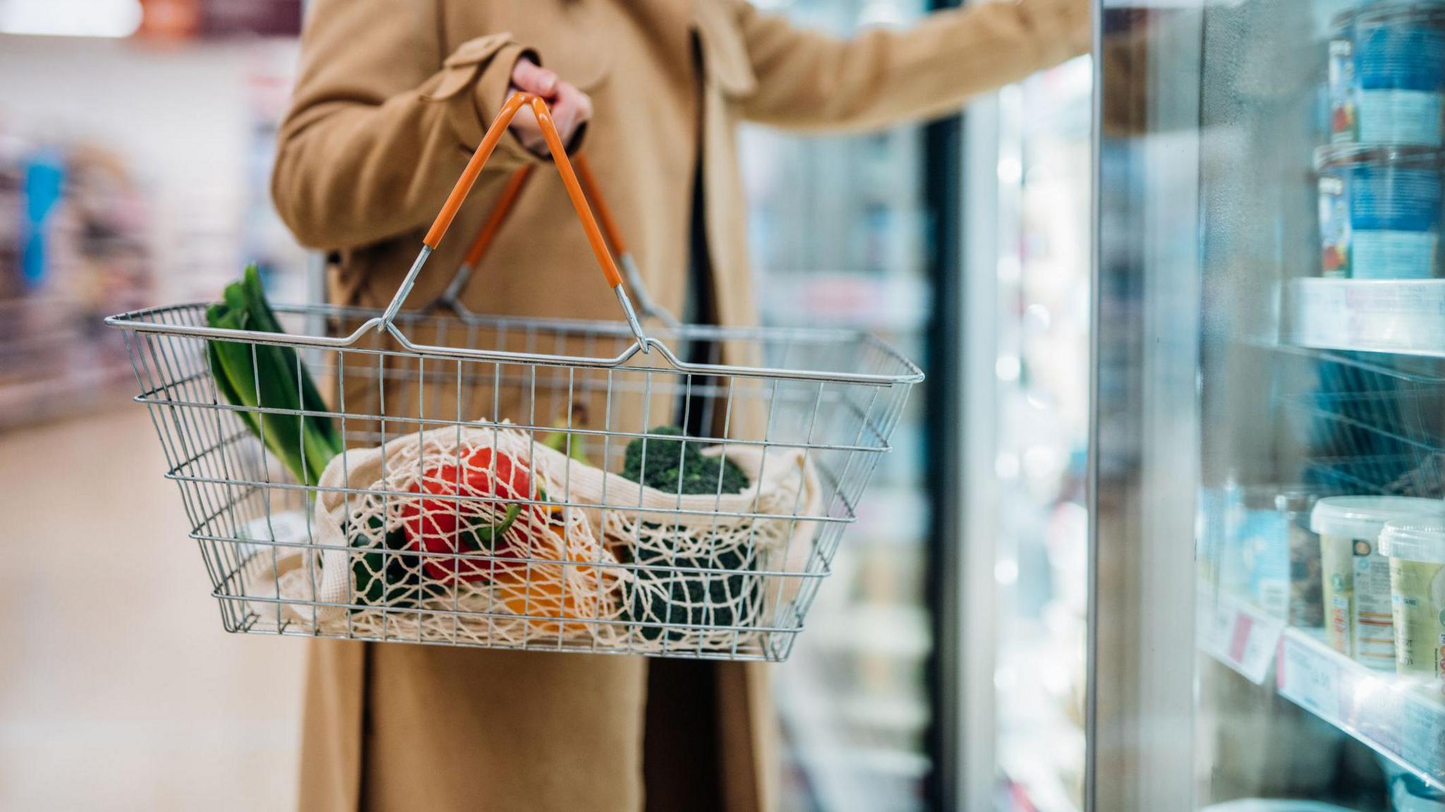 Woman in beige coat shopping near freezer section of supermarket adding stuff to basket