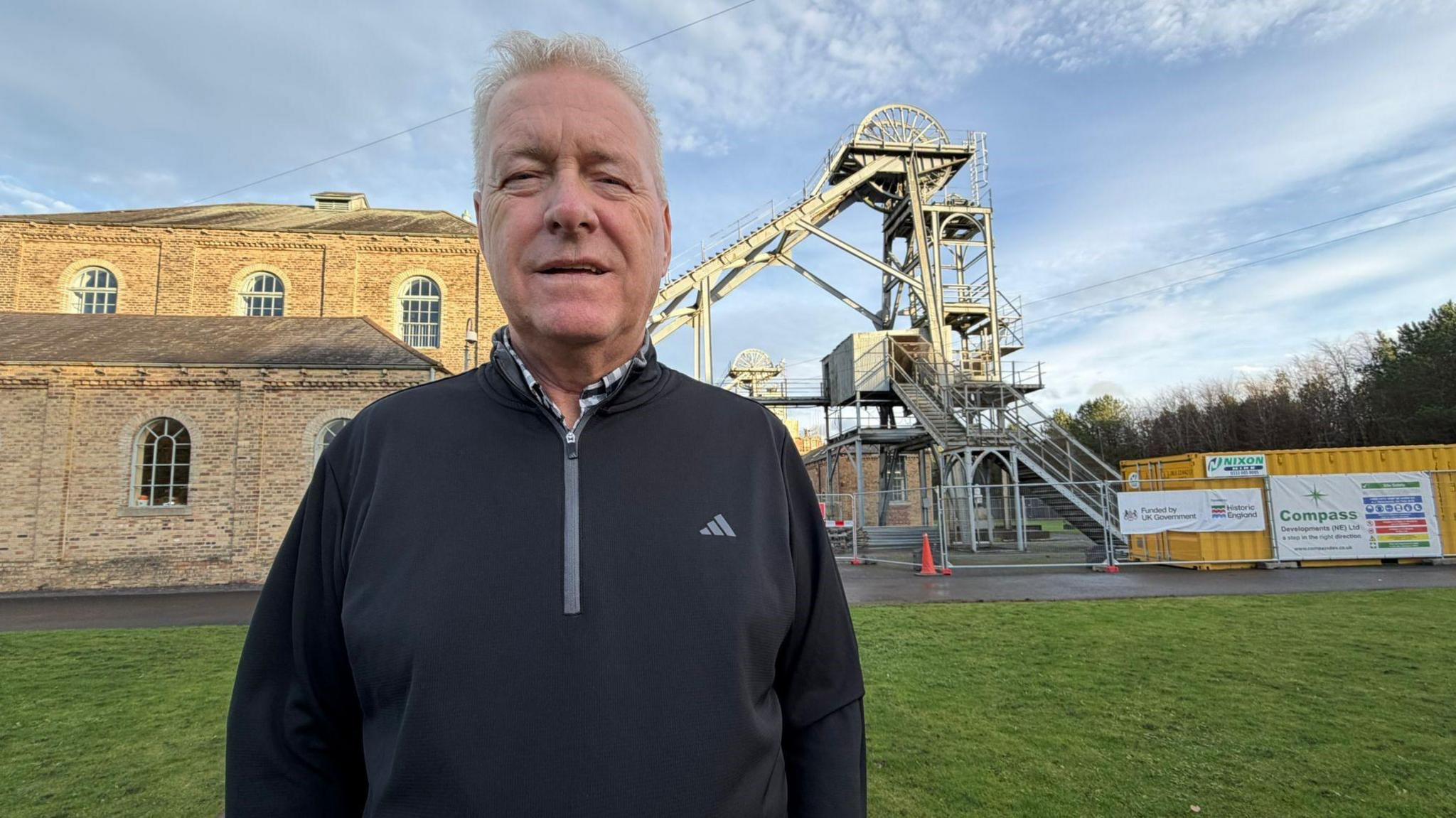 Ian Lavery looking into the camera and smiling. He is standing in front of Woodhorn Colliery which is a stone building with an old bit of mining equipment with is a large metal machine with a wheel on top outside. He has grey short hair  and is wearing a black pullover sports top with a shirt underneath.
